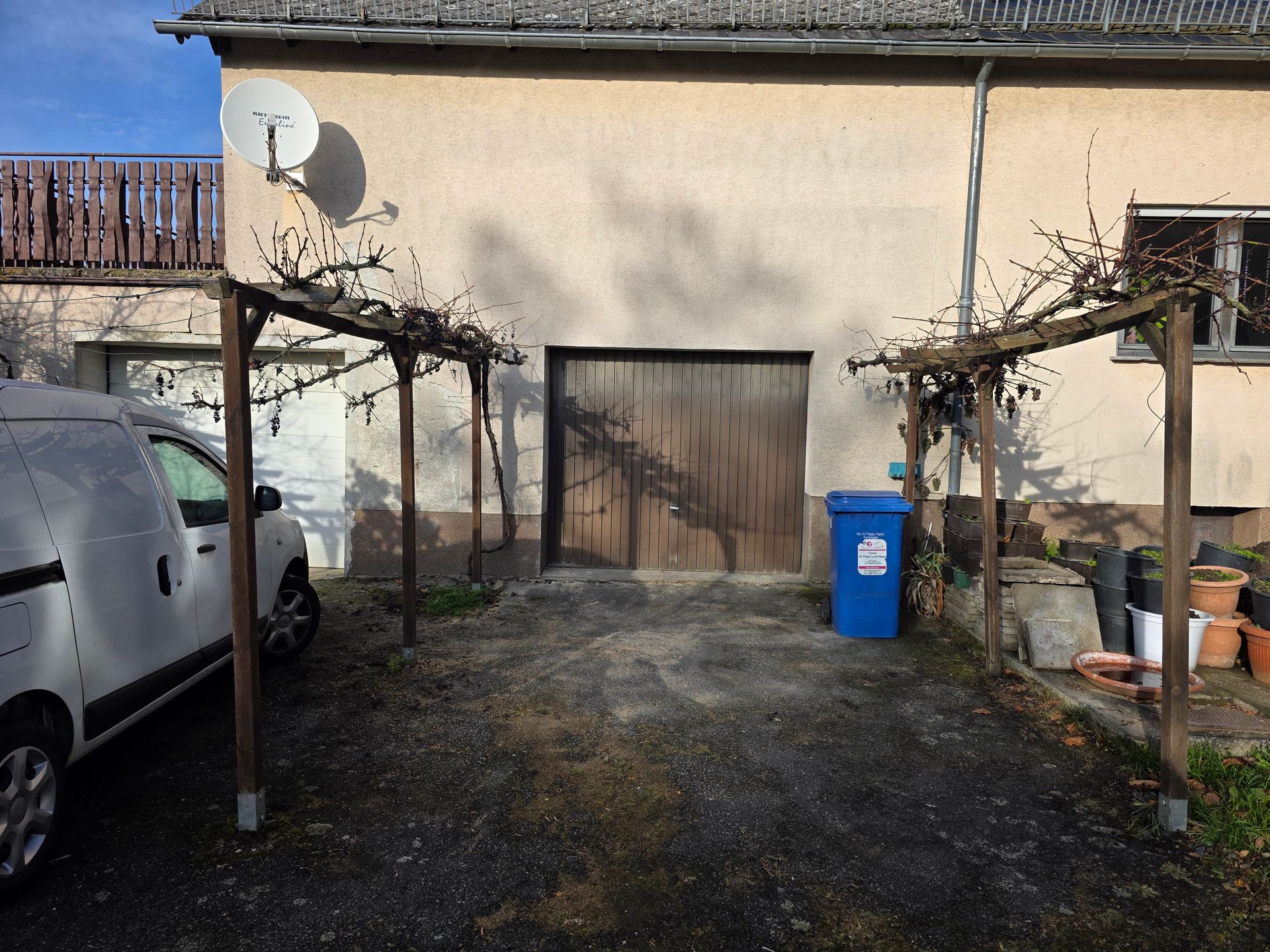 Auffahrt neben einem beigefarbenen Haus mit einem weißen Lieferwagen, einer blauen Mülltonne und zwei hölzernen Rankgittern über einem Kieshof
