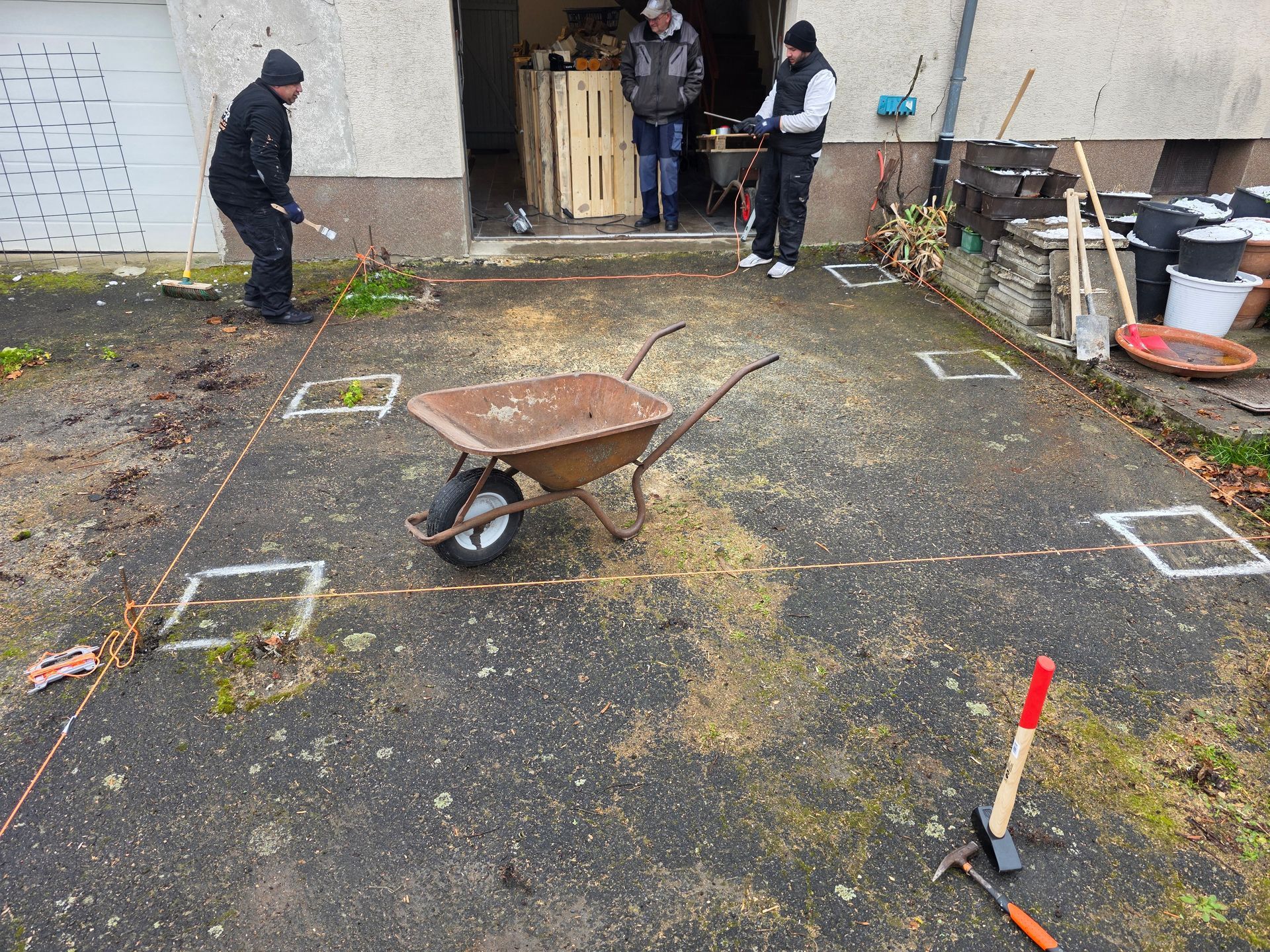 Männer arbeiten auf einer Einfahrt; Schubkarre, Werkzeug und markierte Quadrate sind zu sehen.