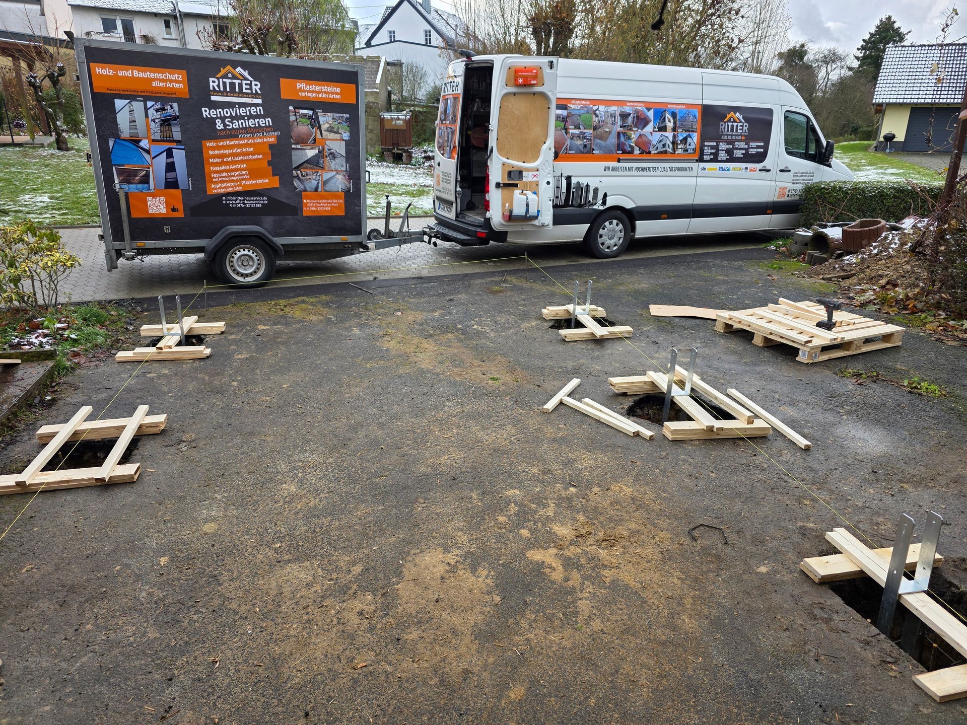 Baustelle mit Anhänger und Lieferwagen, Holzschalungen und einer Palette auf grauem Asphaltbelag.
