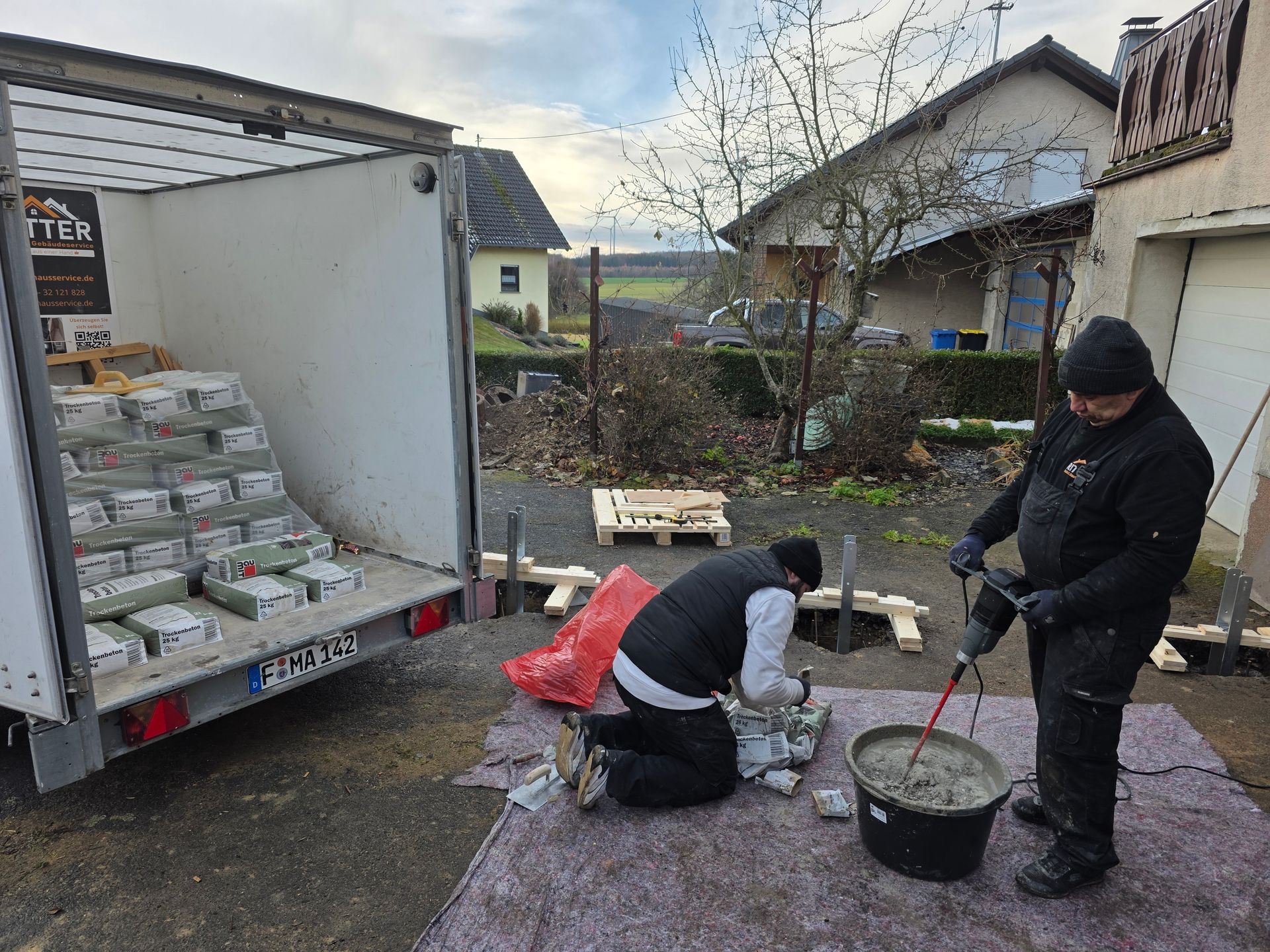 Zwei Personen mischen im Freien Beton neben einem Anhänger, der mit Baumaterialien beladen ist.