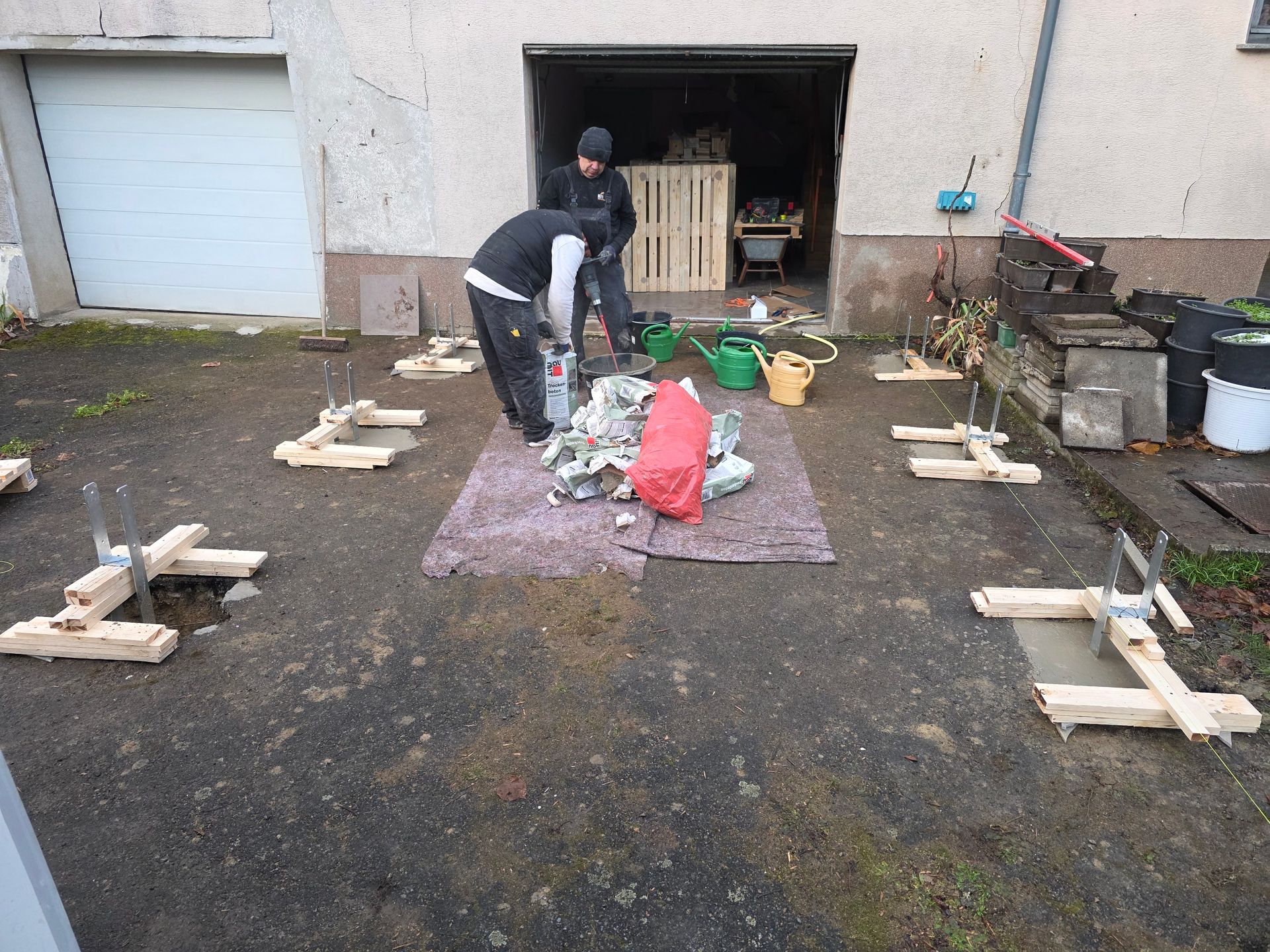 Zwei Personen arbeiten im Freien an einem Projekt, umgeben von Holzständern und Baumaterialien vor einer Garage.