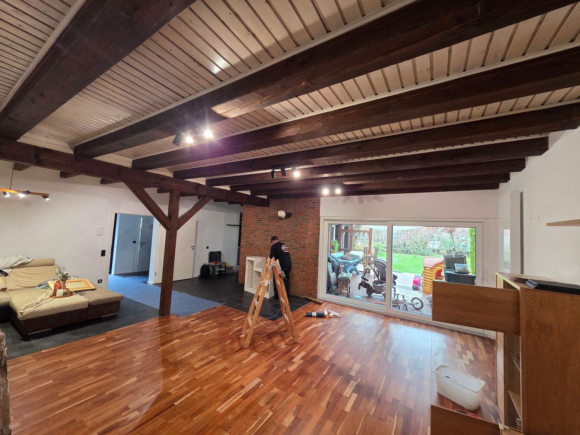 Wohnzimmer mit Holzbalken und Holzboden, Person in der Nähe einer Backsteinwand und Glasschiebetür zum Garten.