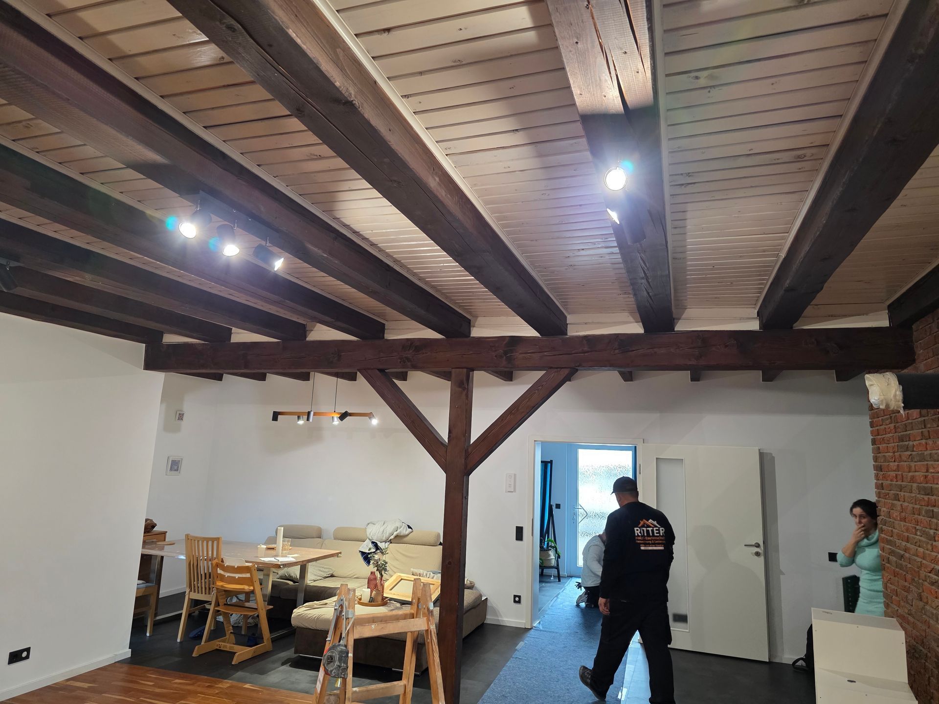 Holzbalkendecke und freiliegende Stützpfeiler in einem Raum mit Menschen und Möbeln.