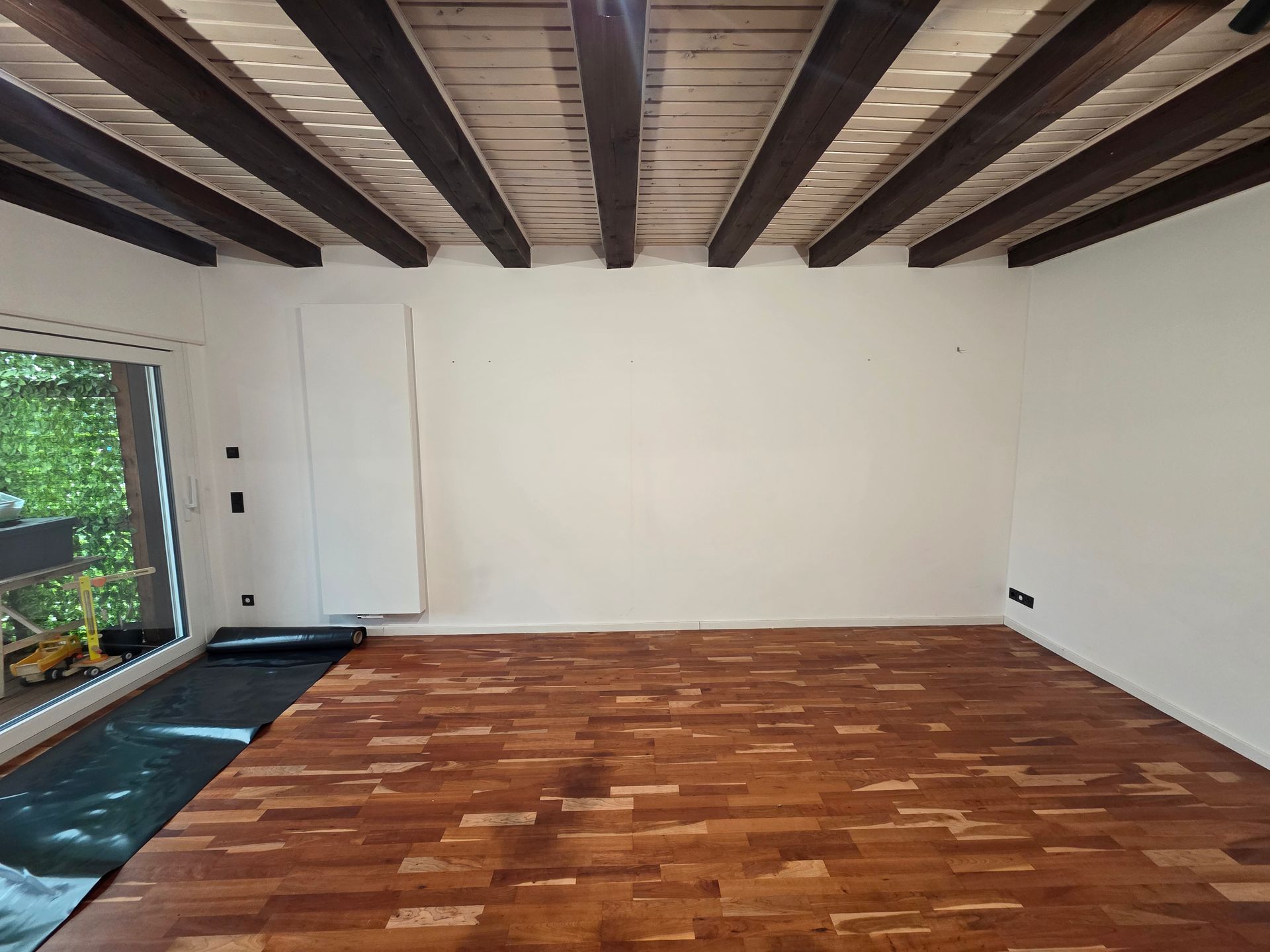 Leerer Raum mit Holzboden und Deckenbalken, einer weißen Wand und einem großen Fenster.