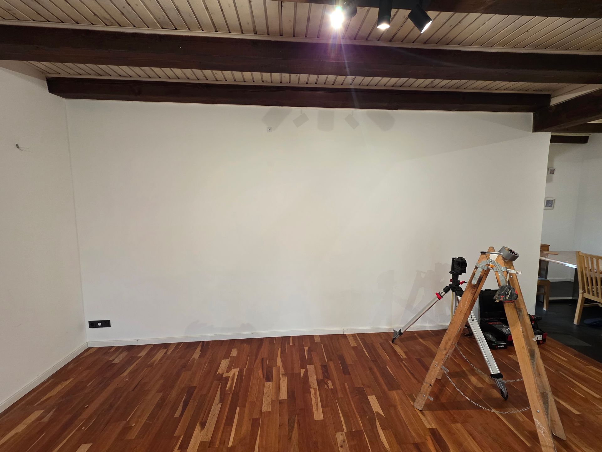 Eine leere weiße Wand in einem Raum mit Holzboden und sichtbaren Holzbalken. Ein Stativ steht vor der Wand.