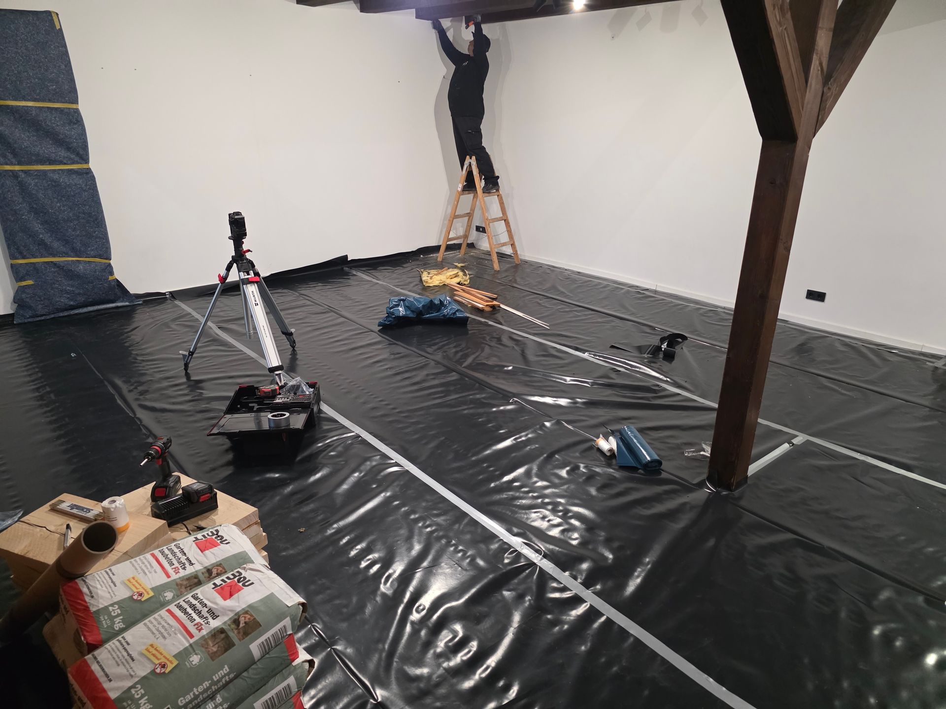 Eine Person arbeitet auf einer Leiter an Deckenbalken in einem Raum mit schwarzem Bodenbelag, Werkzeugen und Dämmmaterial.