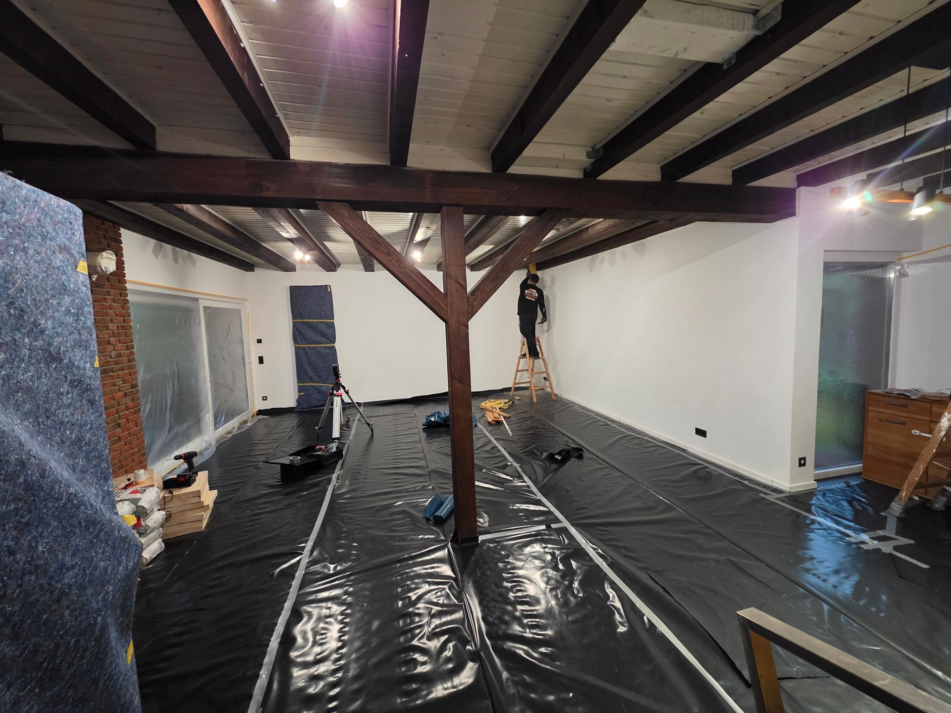 Ein Raum mit Holzbalken, einem zentralen Stützpfosten und einer Person, die eine weiße Wand streicht.