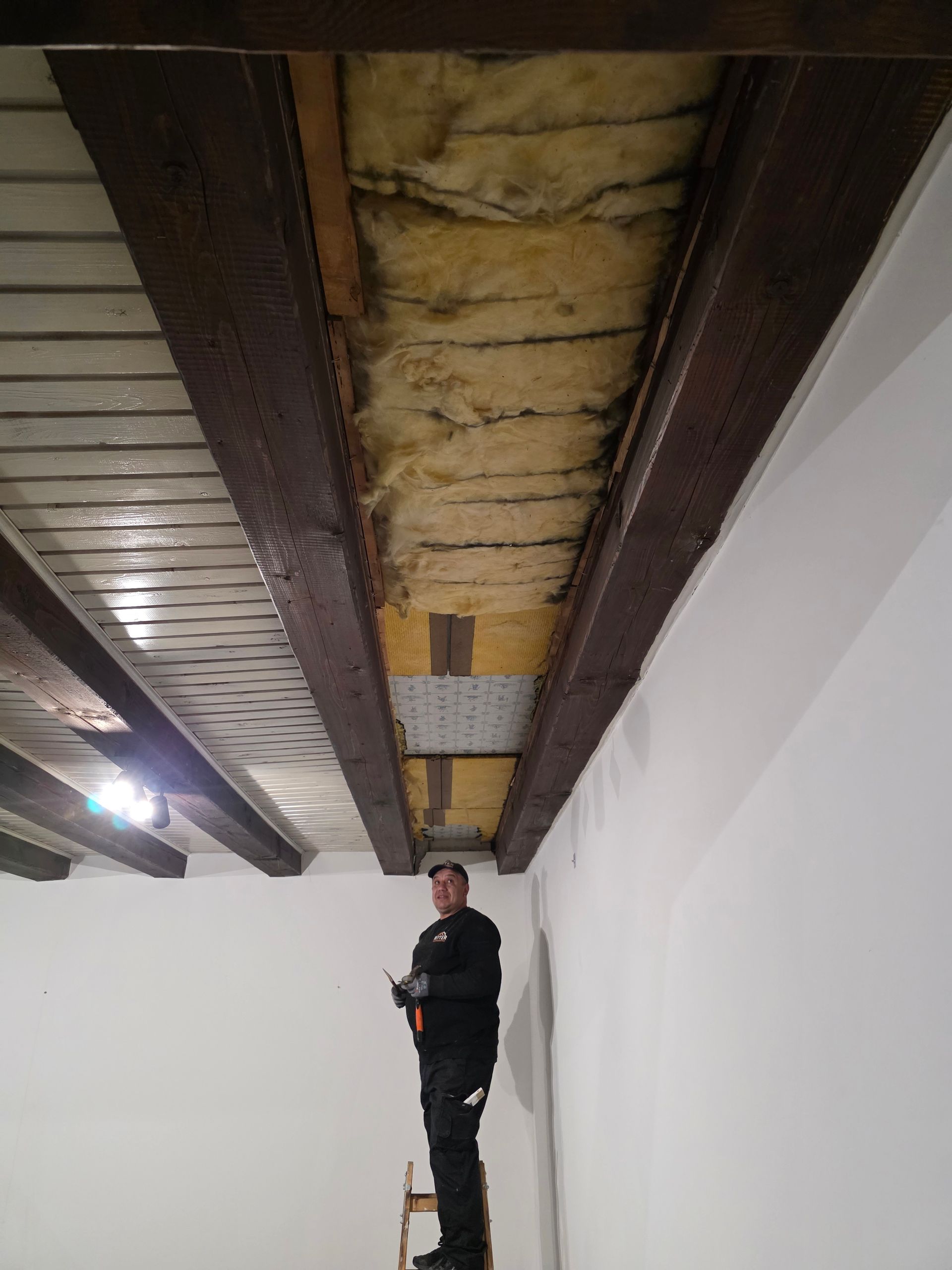 Ein Mann auf einer Leiter untersucht die isolierten Deckenbalken. Weiße Wände und eine metallverkleidete Decke.