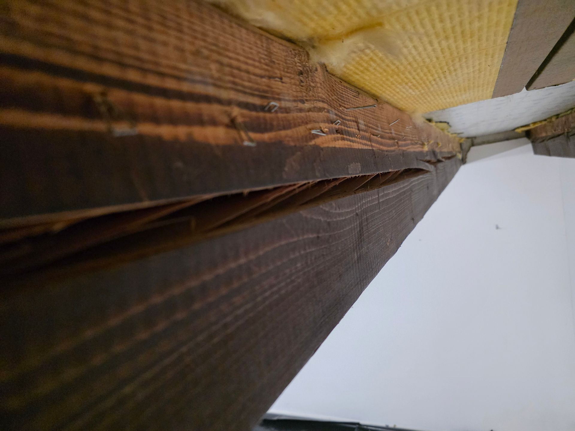 Nahaufnahme eines beschädigten, dunklen Holzbalkens; ein Riss legt die inneren Holzfasern frei. Gelbe Dämmung ist sichtbar.