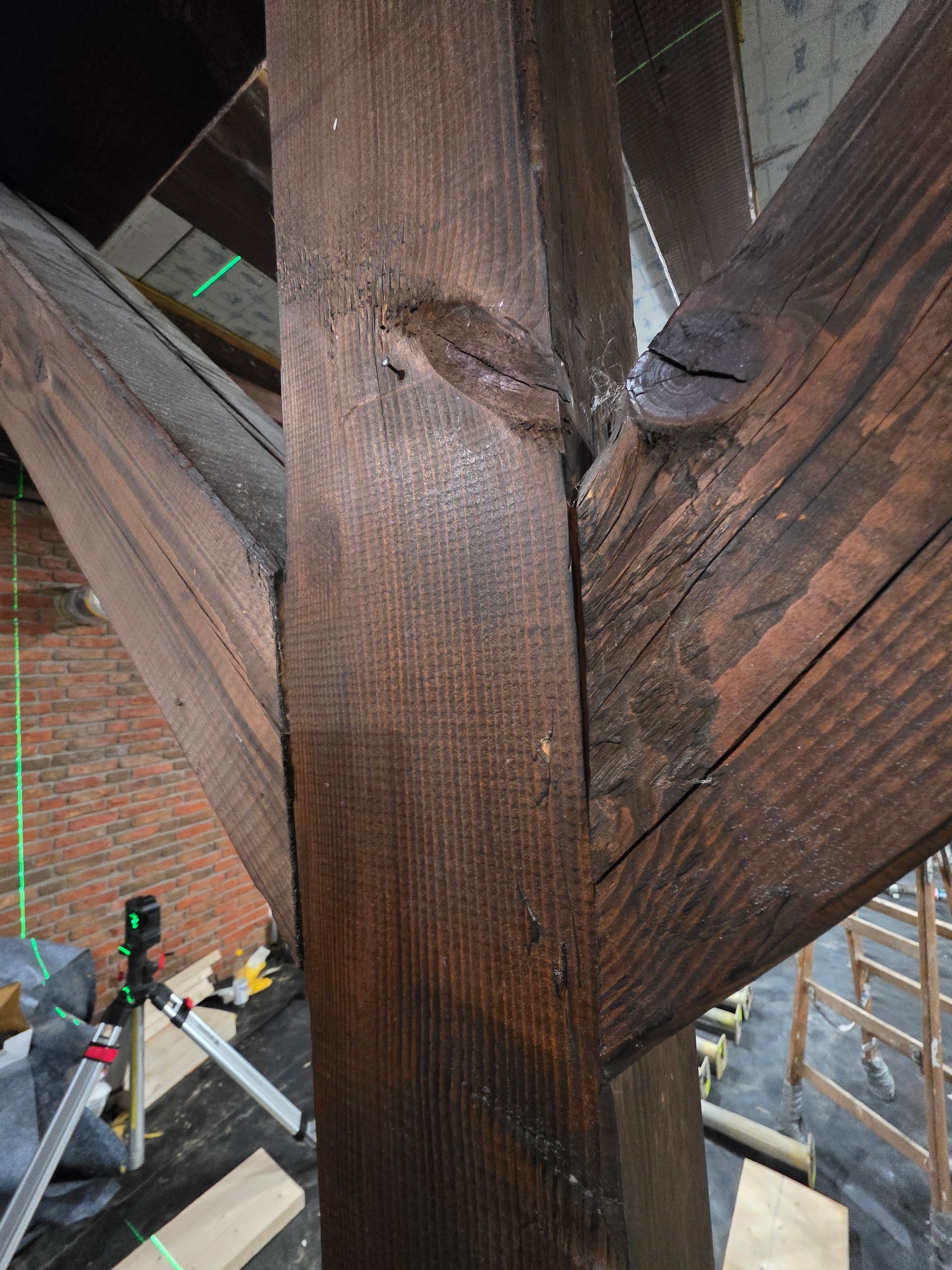 Dunkler Holzbalken und -streben auf einem Dachboden, dahinter sind eine Ziegelwand und Bauwerkzeuge zu sehen.