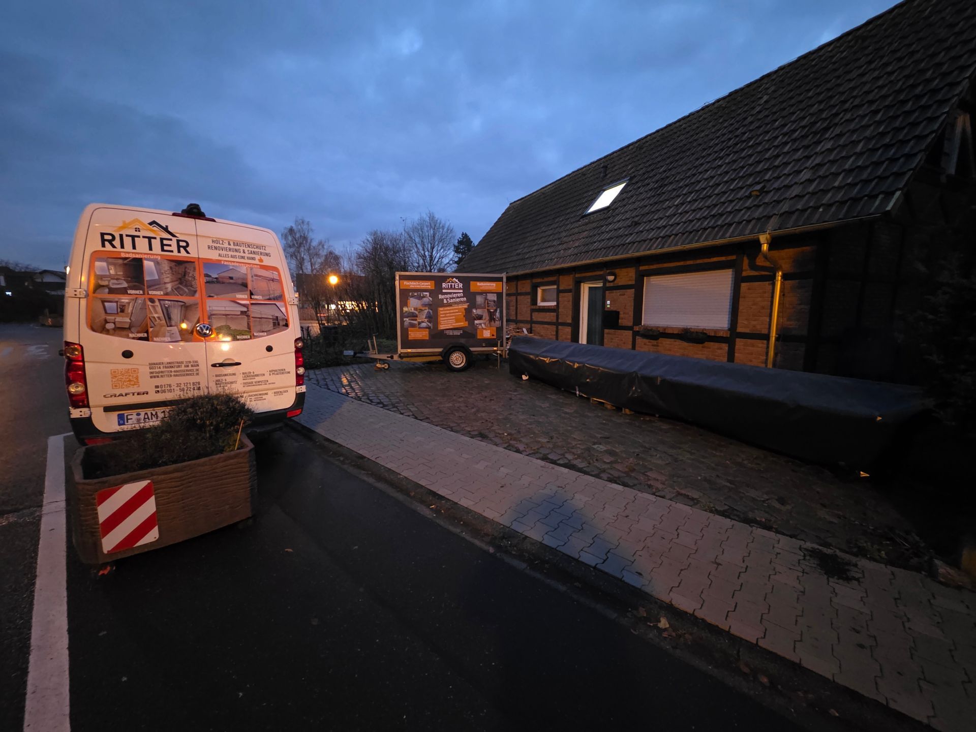 Ein Lieferwagen parkt vor einem Gebäude. Ein Anhänger und ein abgedecktes Objekt sind zu sehen.