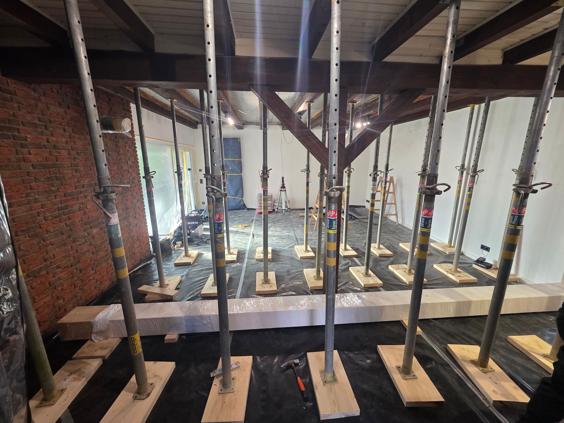 Innenraum mit Baustützen, die eine Decke stützen. Braune Balken und eine Ziegelwand sind sichtbar.