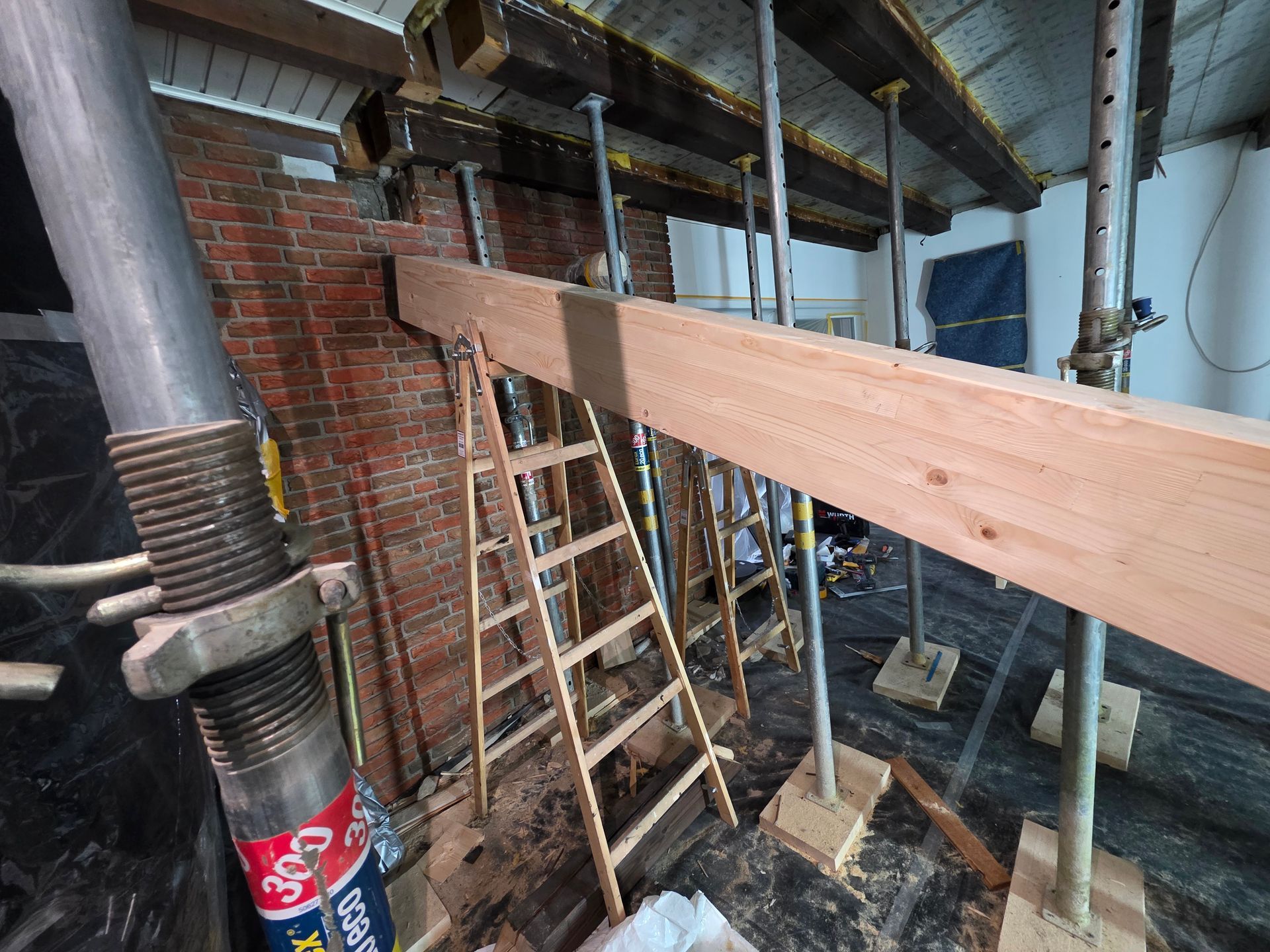 Innenansicht einer Baustelle: freiliegende Ziegelwand, von einem Gerüst getragene Holzbalken, Leiter.