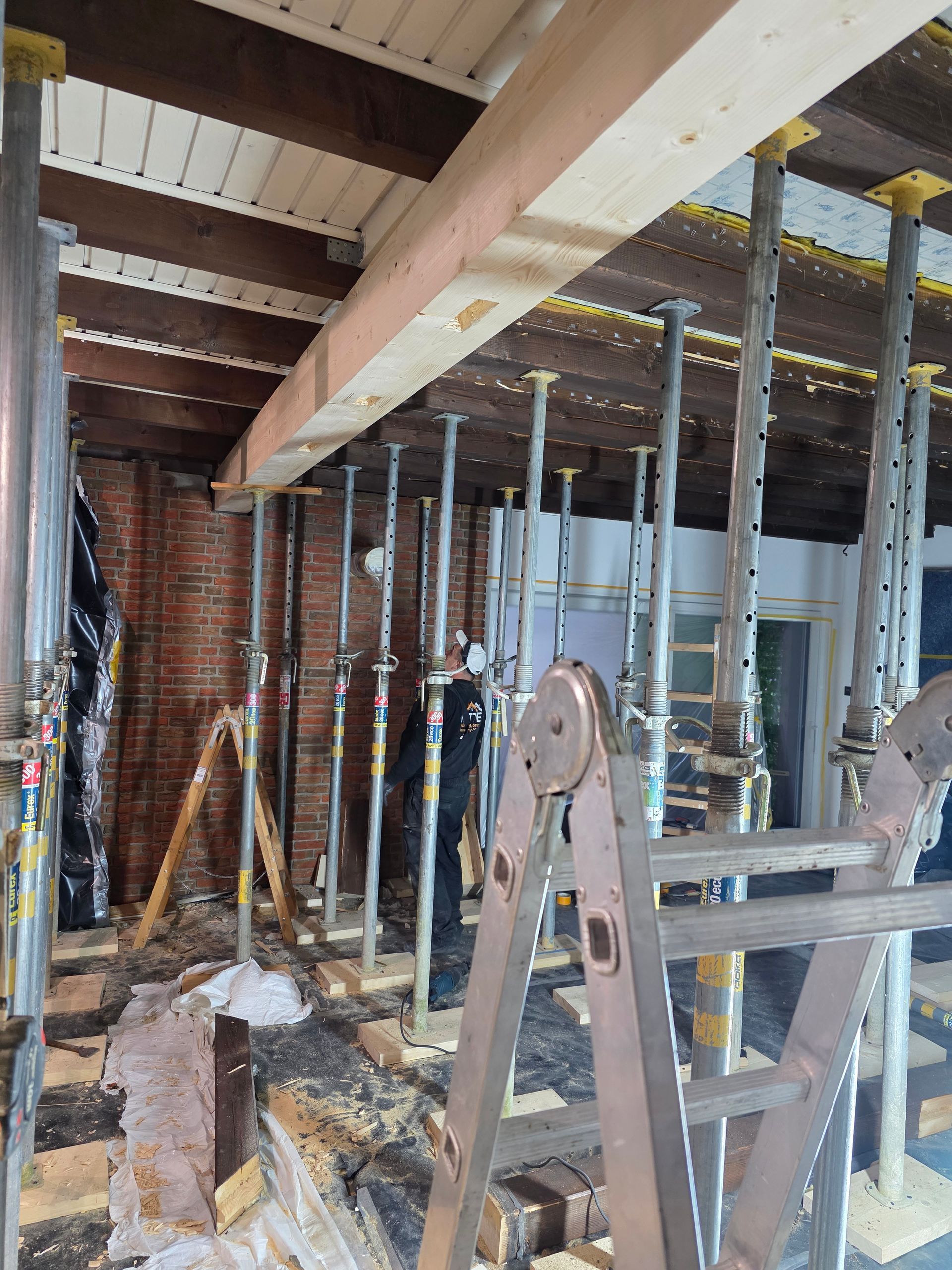 Innenansicht einer Baustelle. Holzbalken werden von Metallpfosten getragen. Ein Arbeiter steht daneben.