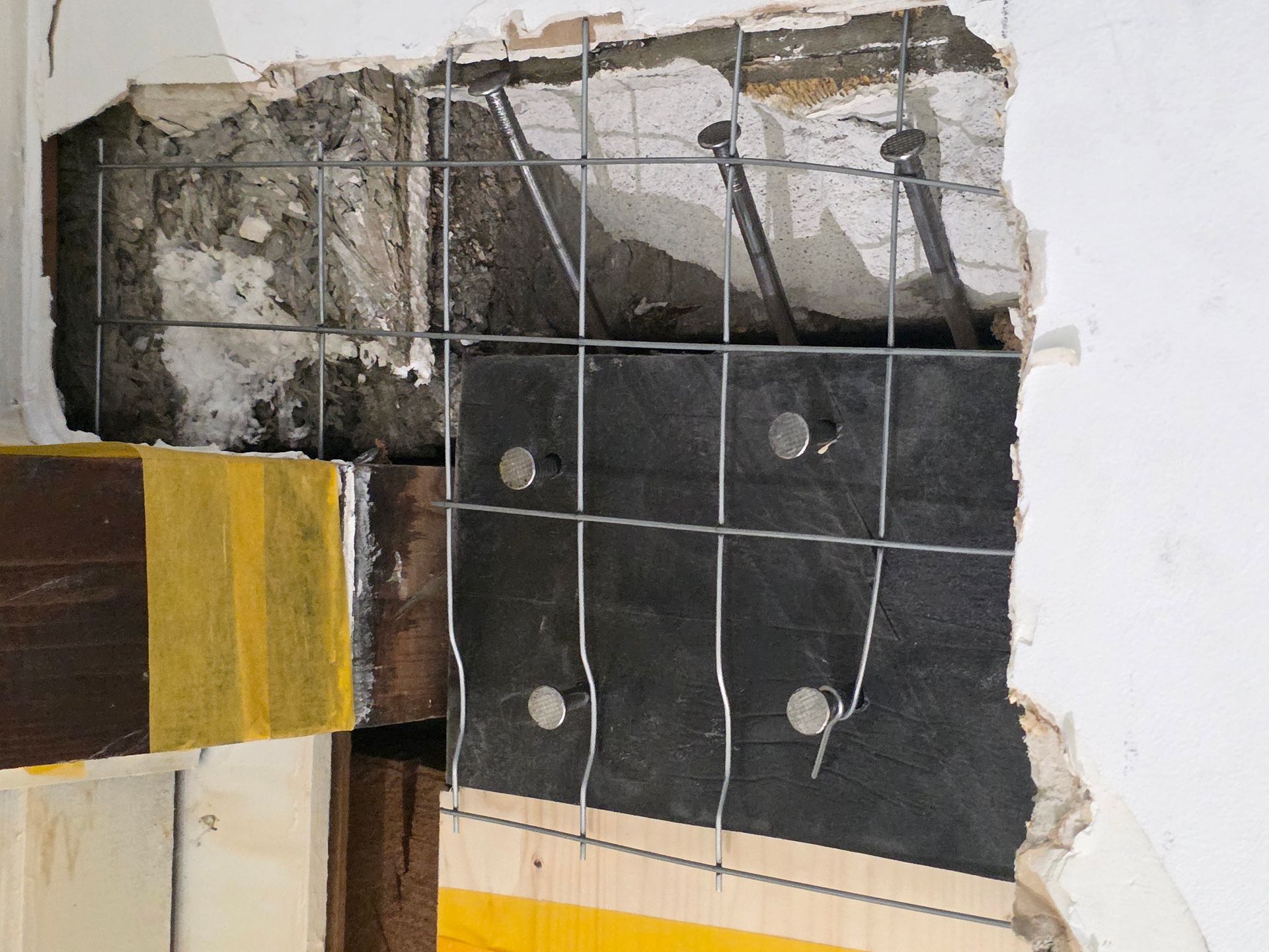 Wandreparatur: Metallgitterarmierung, Sichtbeton, Holzbalken und schwarze wasserdichte Membran.