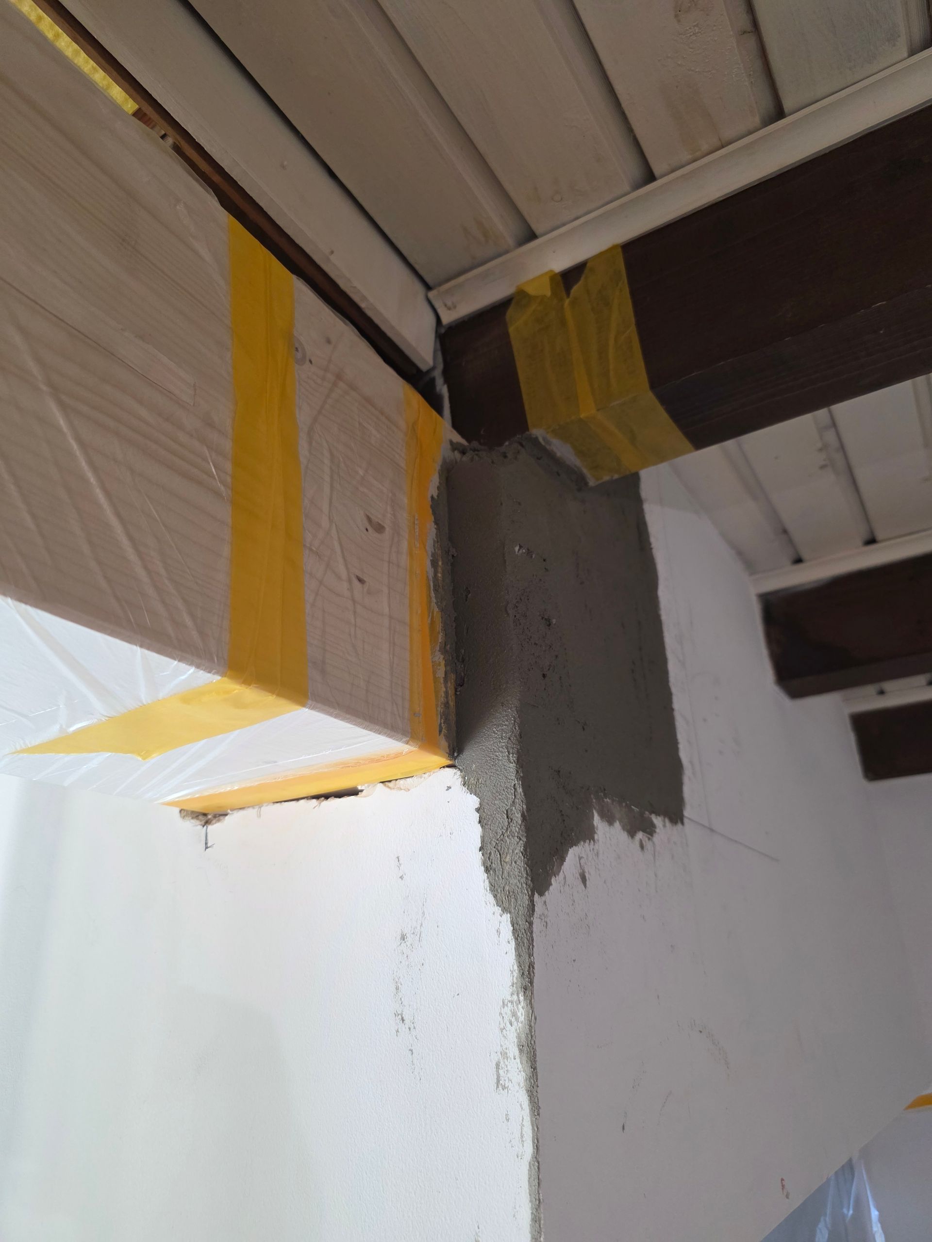 Eine Zimmerecke mit einer Betonplatte an der Wand, die Holzbalken trägt. Gelbes Absperrband umgibt eine Plastikplane.