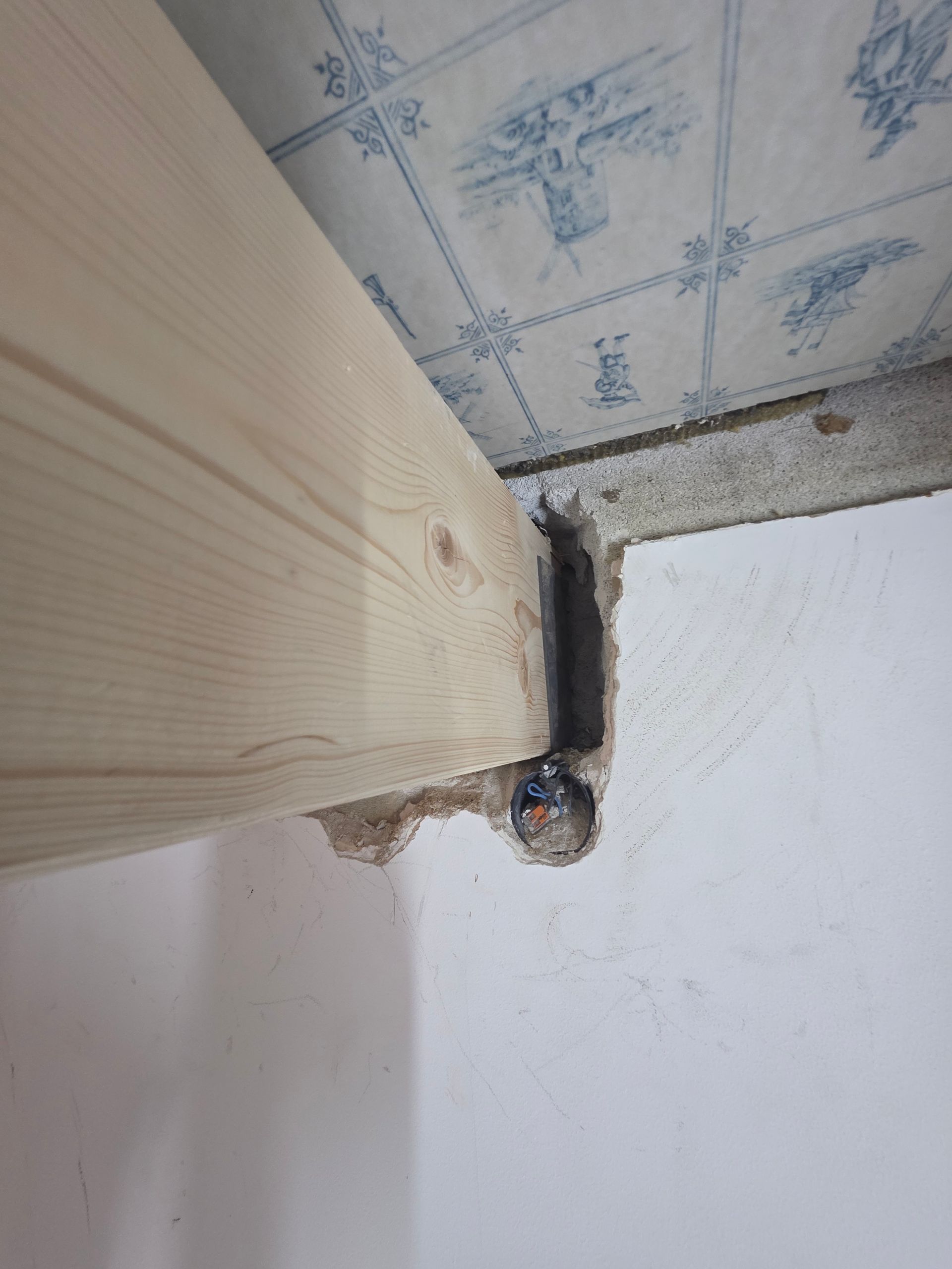 Ein Holzbalken ist an einer Betonwand befestigt, darüber befindet sich eine geflieste Decke und seitlich eine weiße Wand.