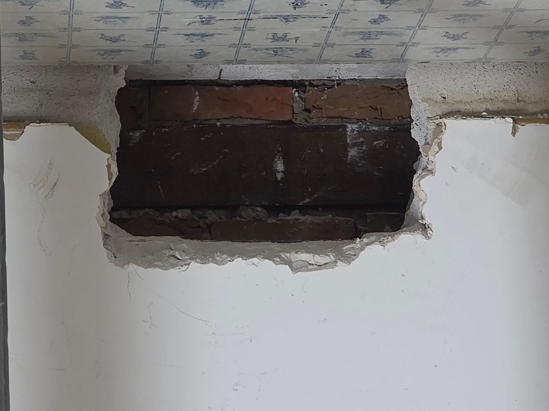 Ein Loch in einer weißen Wand gibt den Blick auf Ziegelstein frei; darüber befinden sich blau gemusterte Deckenplatten.