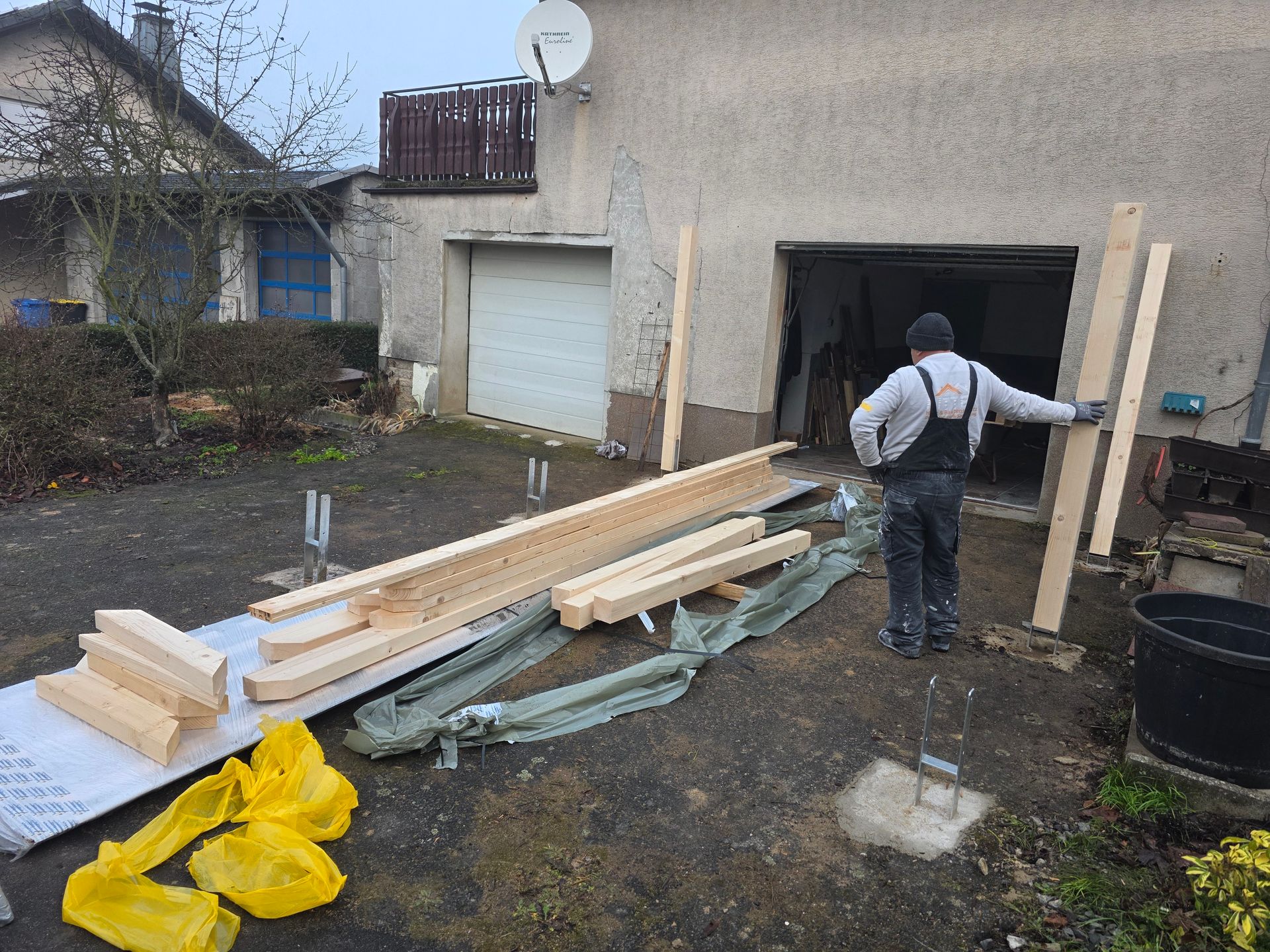 Eine Person steht neben Bauholz vor einer Garage und bereitet sich vermutlich auf Bauarbeiten vor. Bewölkter Tag.
