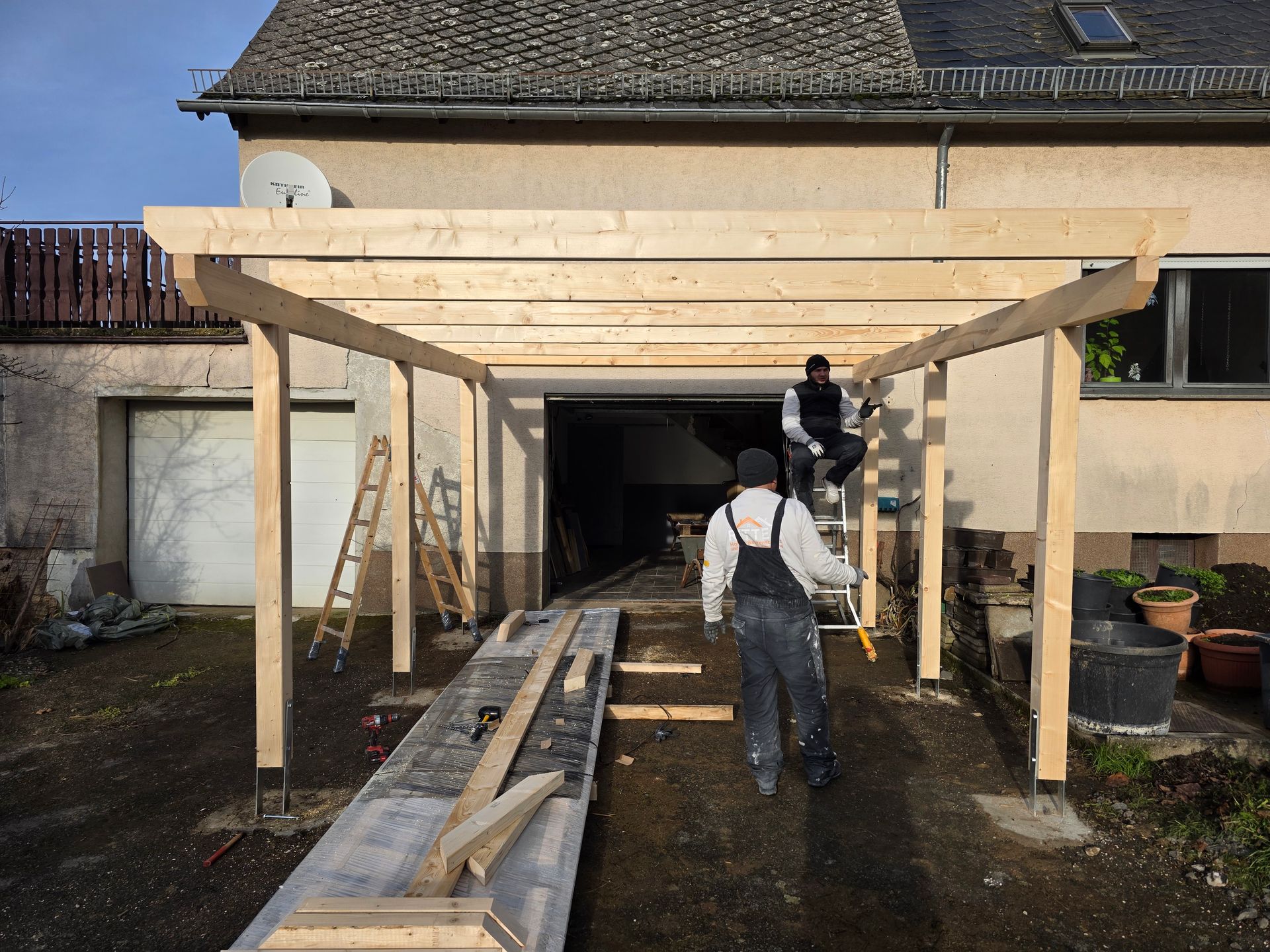 Zwei Personen errichten vor einem Haus eine hölzerne Carportkonstruktion.