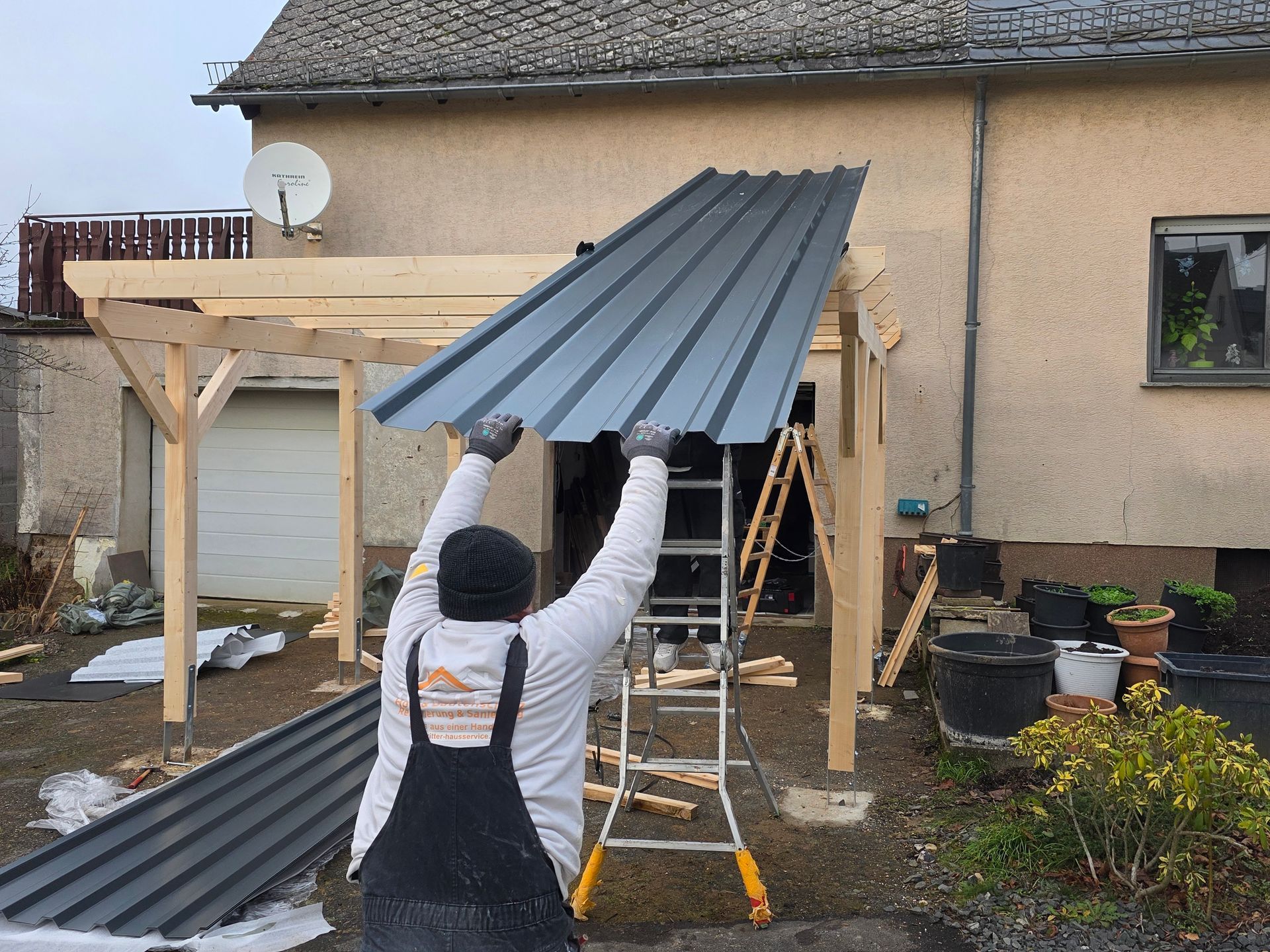 Eine Person montiert eine Metalldachplatte auf einer hölzernen Pergola außerhalb eines Gebäudes.