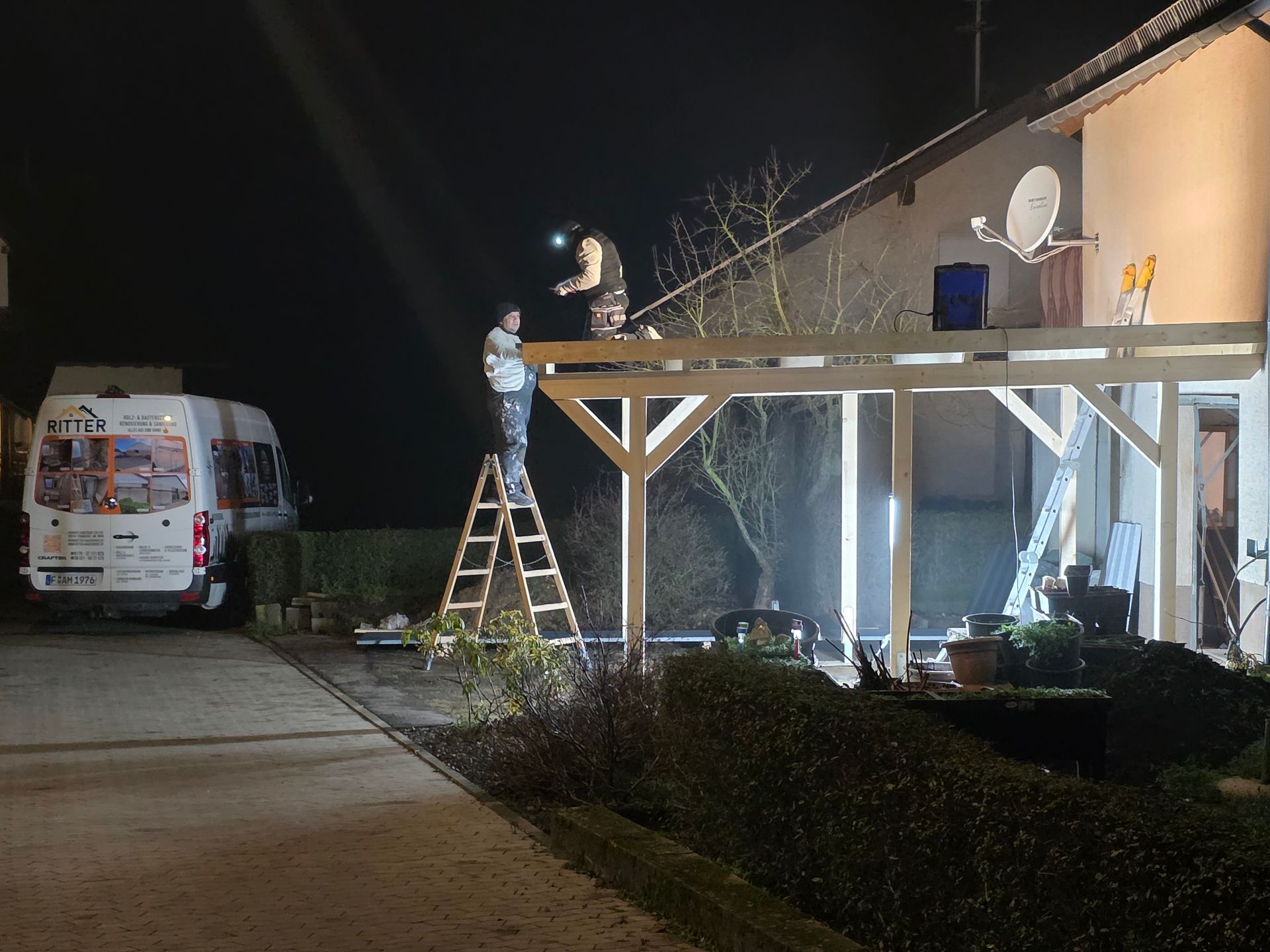 Zwei Personen bauen nachts eine Holzkonstruktion an einem Haus an, eine davon auf einer Leiter. 
