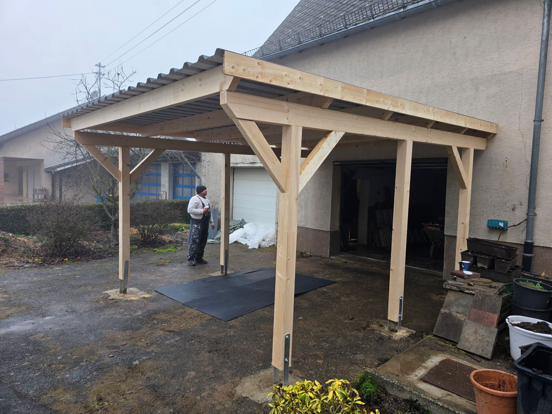 Ein hölzerner Carport ist an ein beiges Gebäude angebaut; eine Person steht darin.