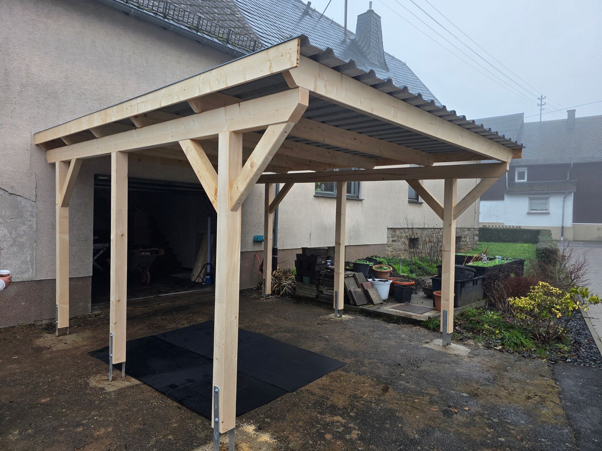 Ein hölzerner Carport, der an eine Garage angebaut ist; ein helles Bauwerk mit einem Dach aus Wellblech.