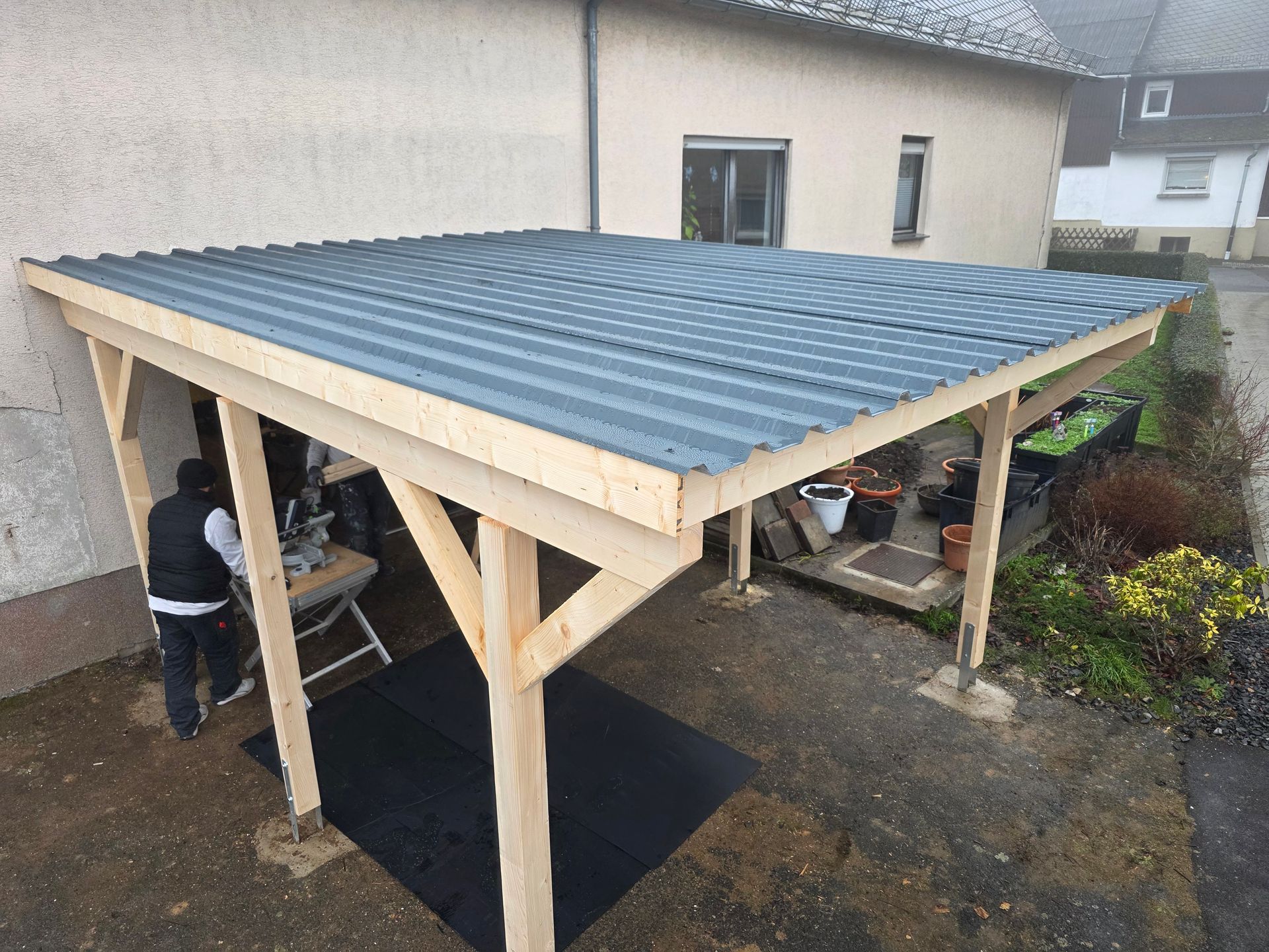 Holzcarport mit einer arbeitenden Person; graues Dach, Betonfundament, in der Nähe eines Gebäudes.