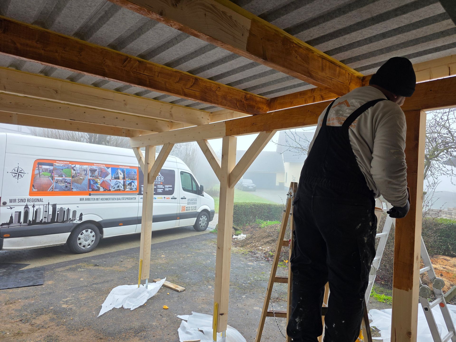 Ein Bauarbeiter streicht auf einer Leiter eine Holzkonstruktion; im Hintergrund ist ein Lieferwagen zu sehen.
