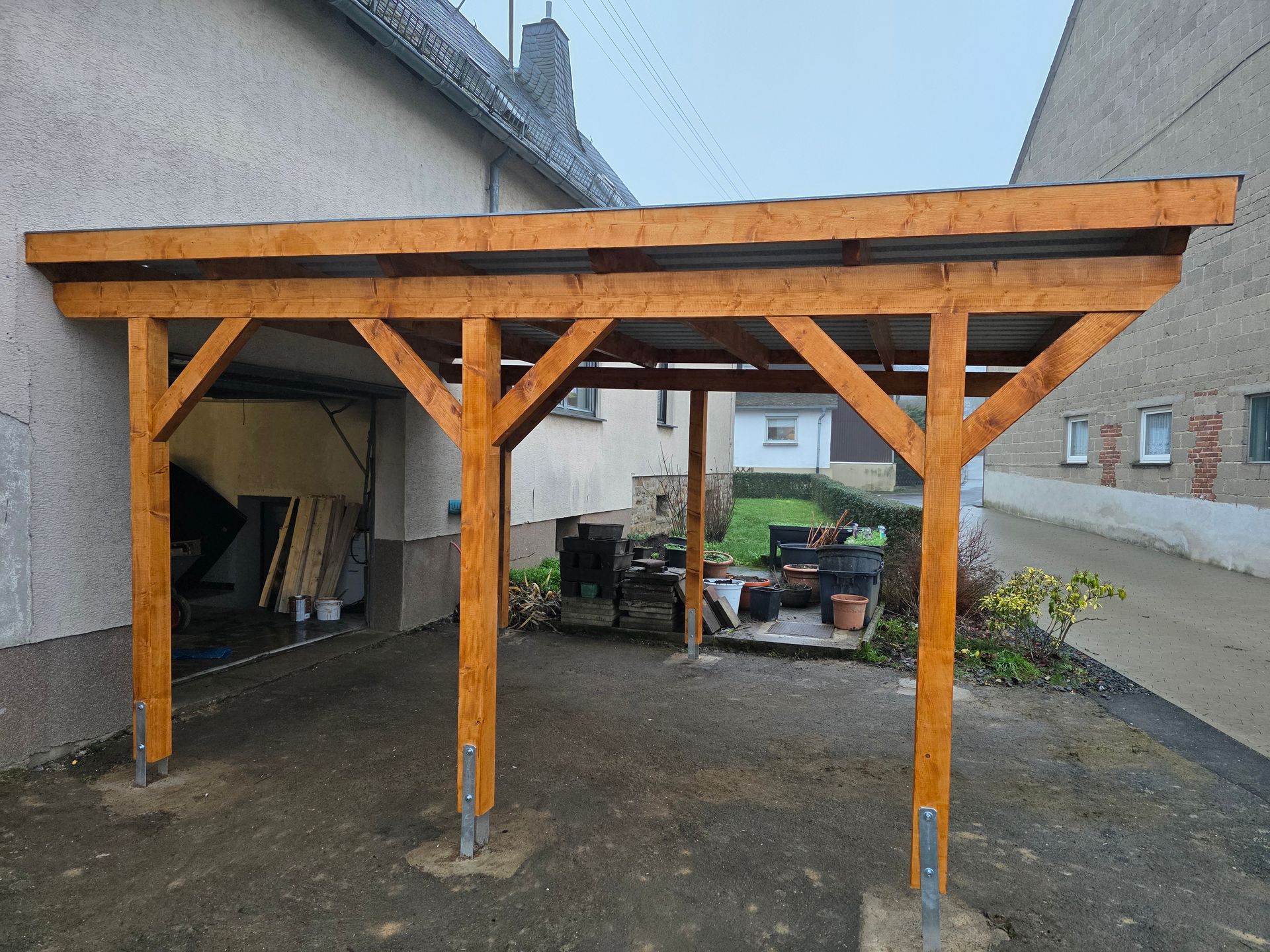 An ein Gebäude angebauter Carport aus Holz, braunes Gestell, graues Dach, im Freien gelegen.