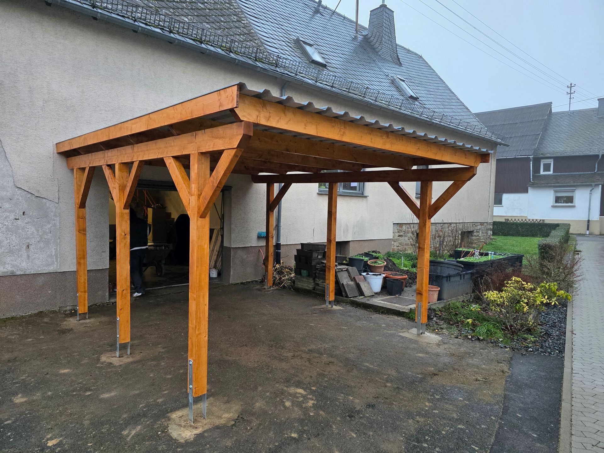 Ein hölzerner Carport, der an ein helles Haus angebaut ist und die Einfahrt überdacht, mit Wellblechdach.