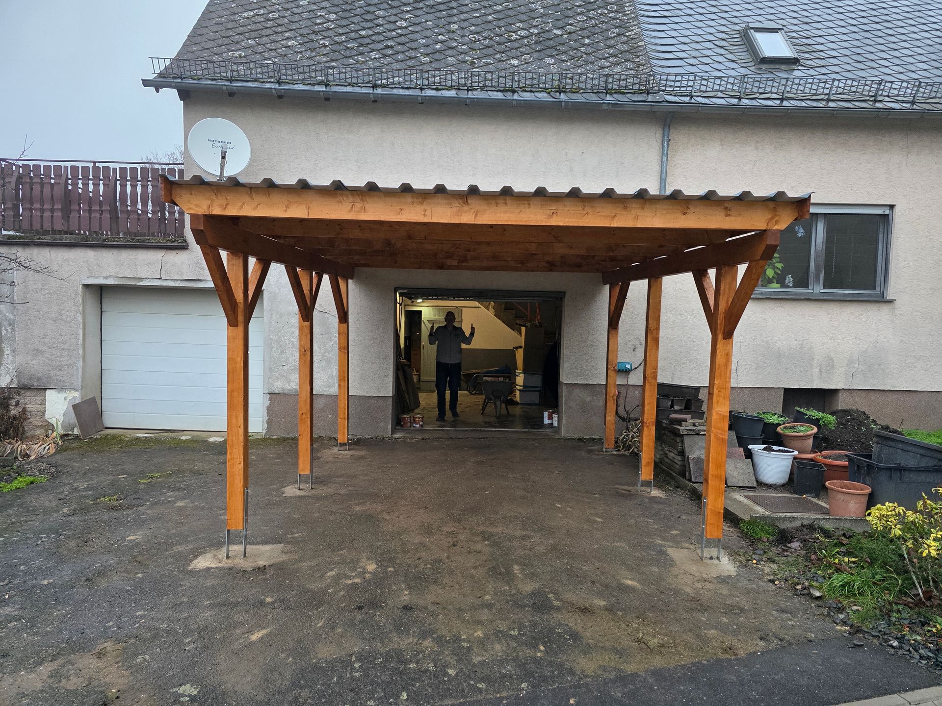 Ein hölzerner Carport ist an ein Haus angebaut, in dessen Türrahmen eine Person steht; Hof und Garage sind von dort aus zu sehen.