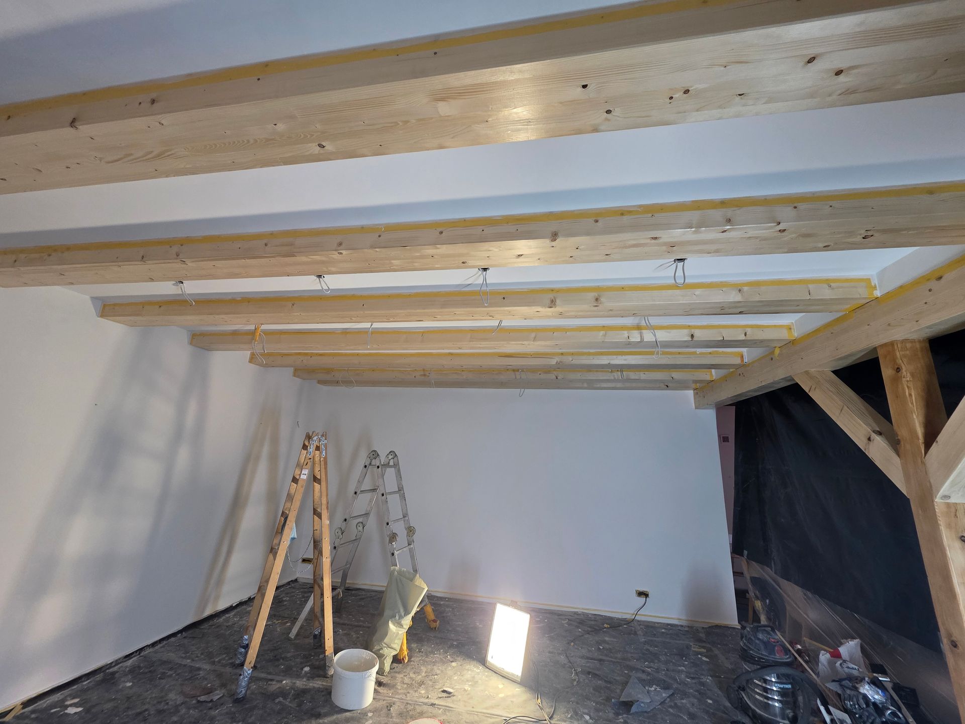 Ein im Bau befindlicher Raum mit freiliegenden Holzbalken, Leitern und weißen Wänden.