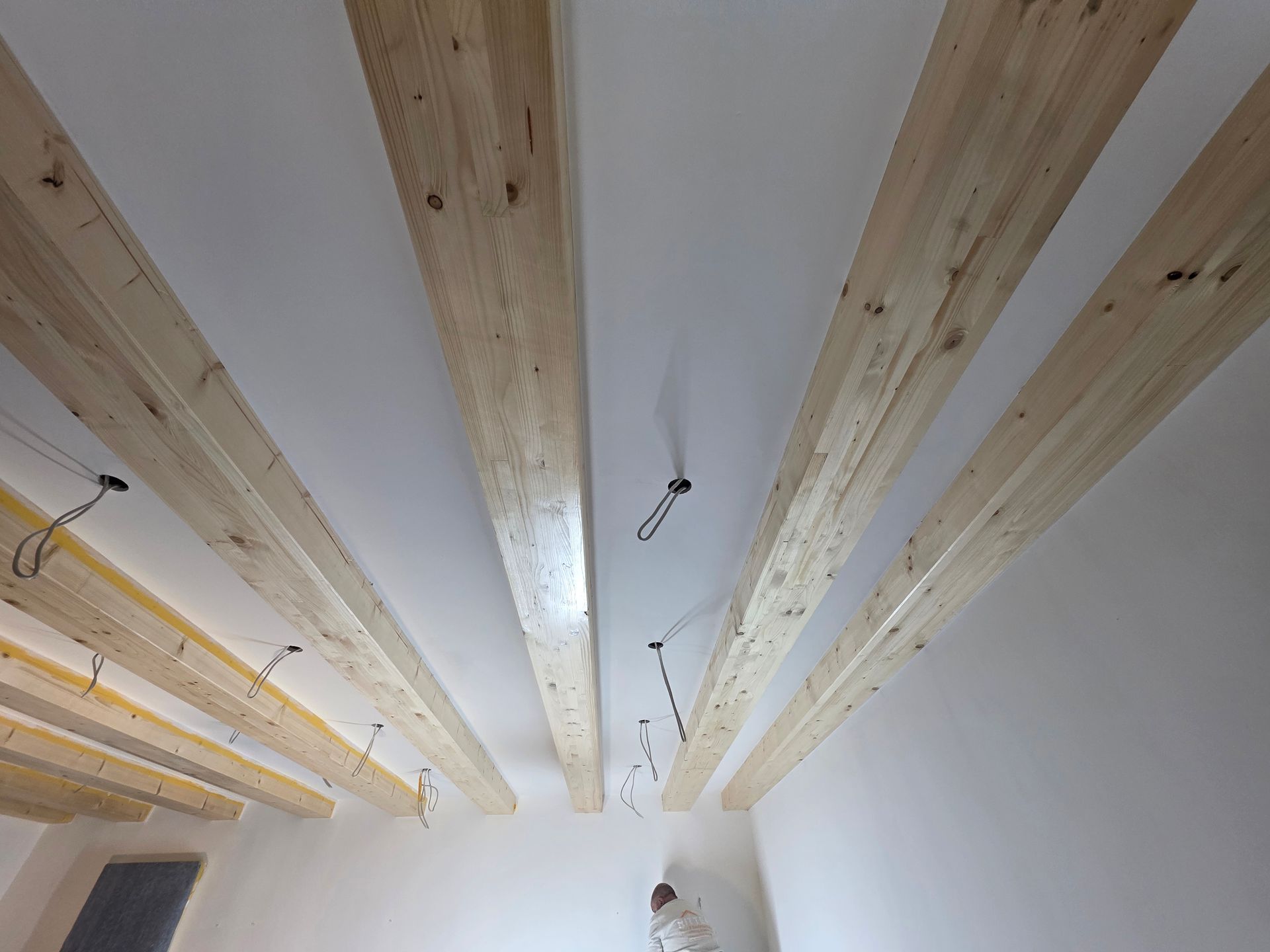 Holzbalken an weißer Decke, herabhängende Stromkabel, ein sichtbares Fenster und eine Person.