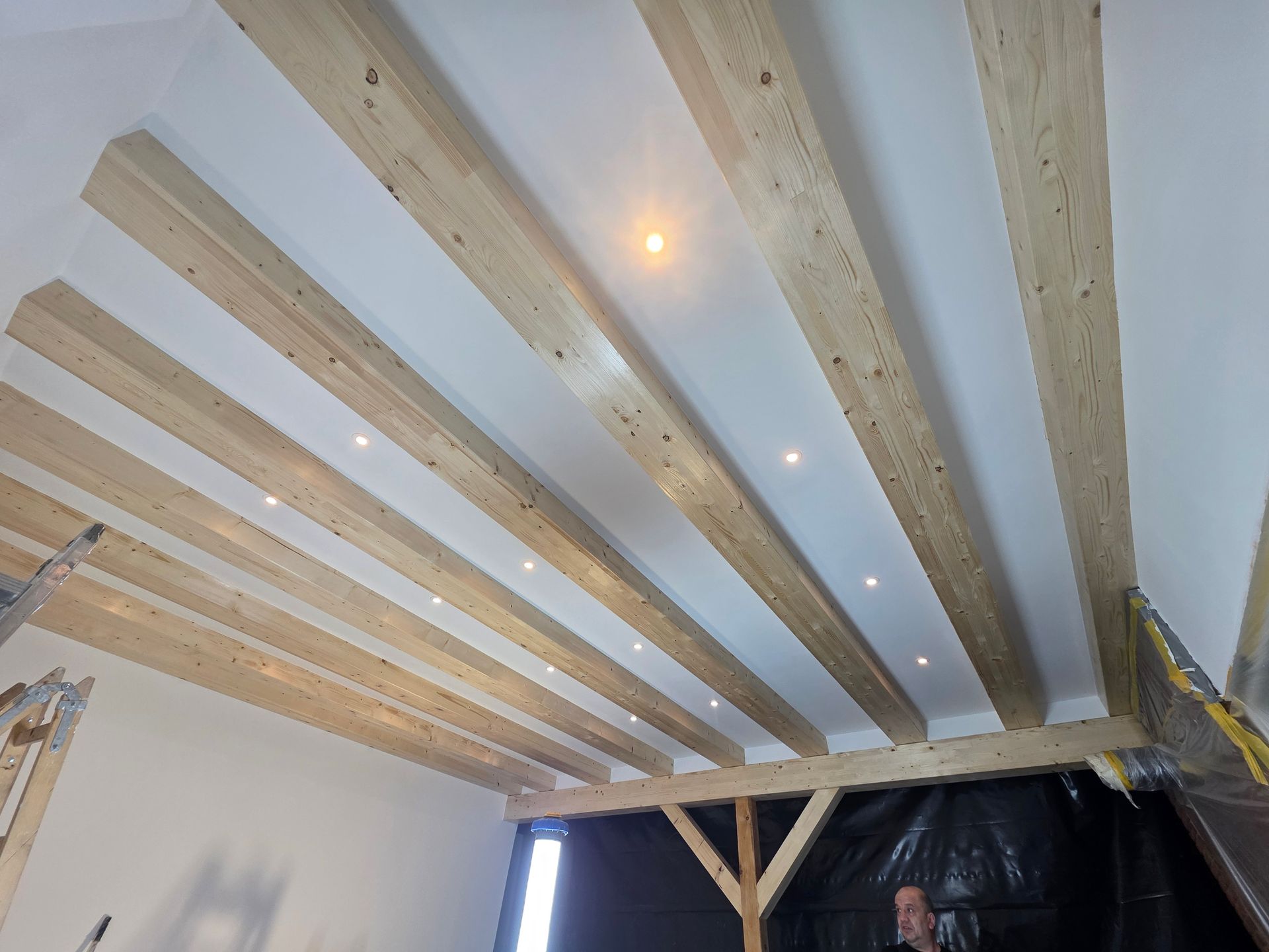 Holzbalken an einer weißen Decke mit Einbaustrahlern. In der unteren rechten Ecke ist eine Person zu sehen.
