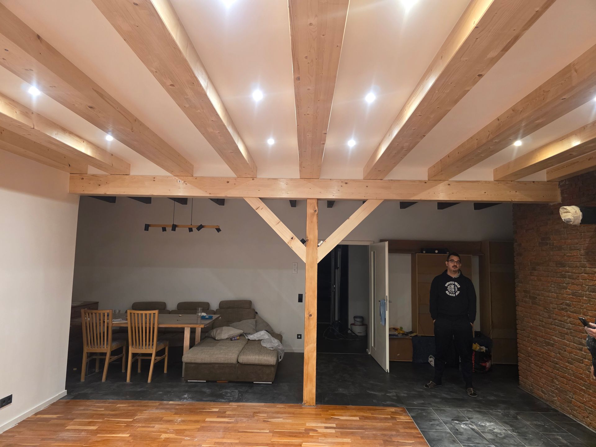 Holzbalkendecke in einem renovierten Wohnraum, Person steht in der Nähe der Tür, Backsteinwand, Essbereich sichtbar.