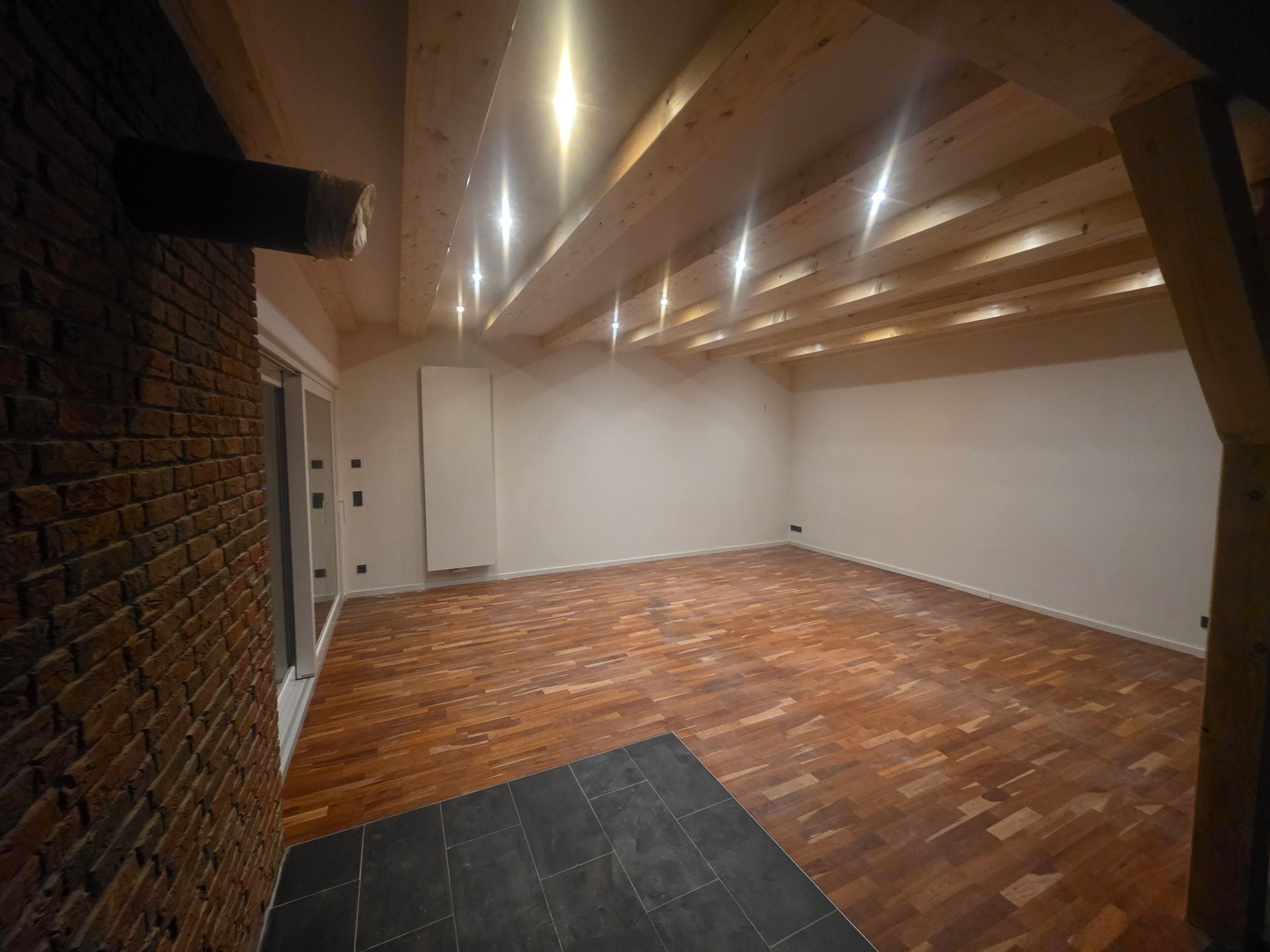 Leerer Raum mit Holzboden, Ziegelwand und freiliegenden Holzbalken an der Decke.