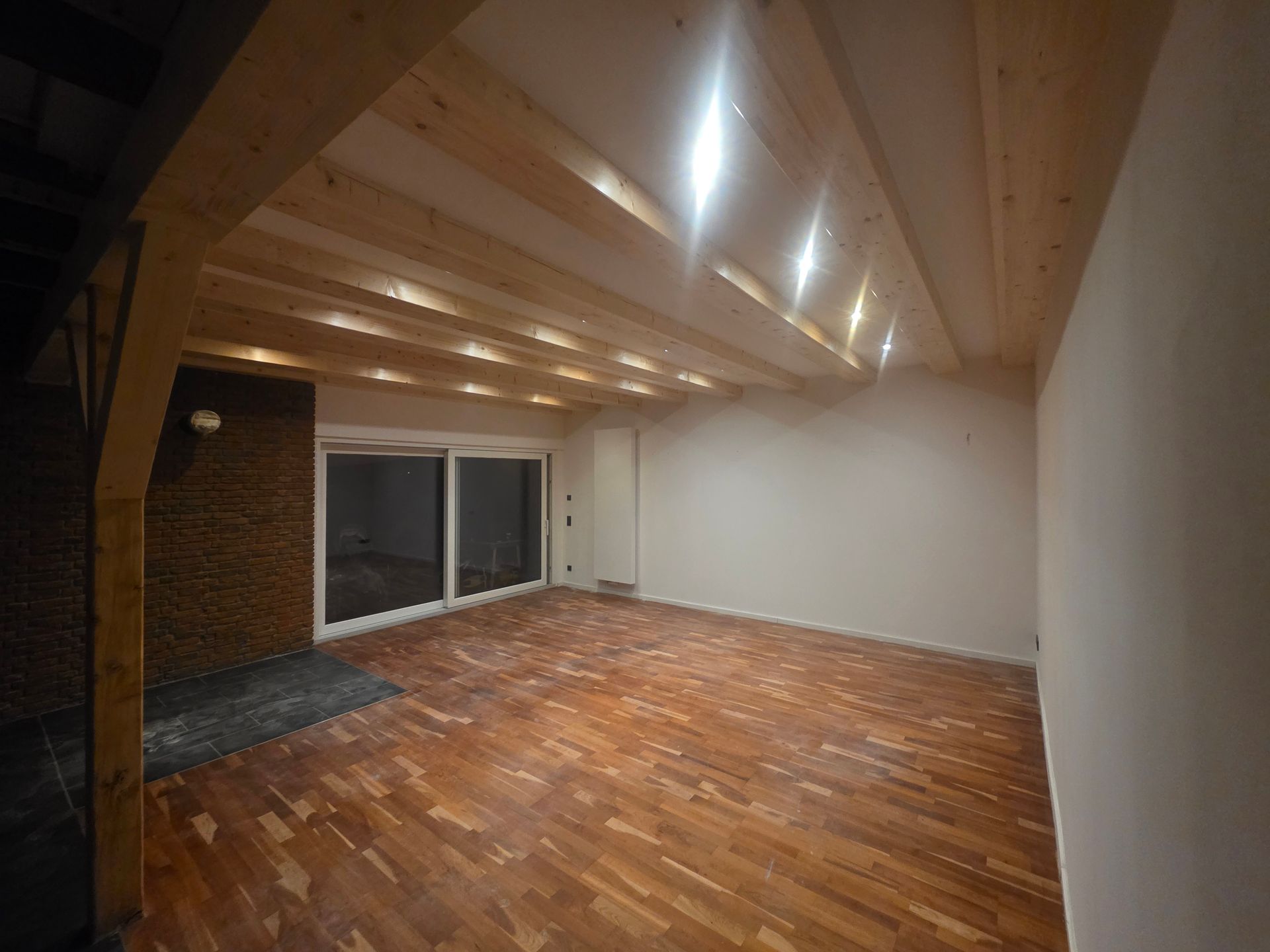 Leerer Raum mit Holzbalken, Glasschiebetüren, Holzboden und Ziegelwand.