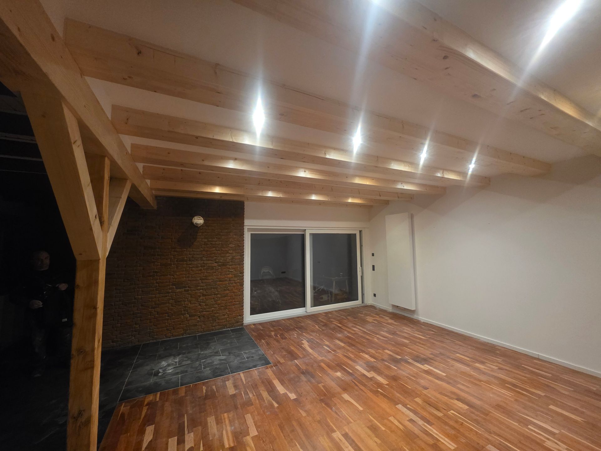 Zimmer mit Holzboden, sichtbaren Holzbalken, einer Glasschiebetür und einer Ziegelwand.