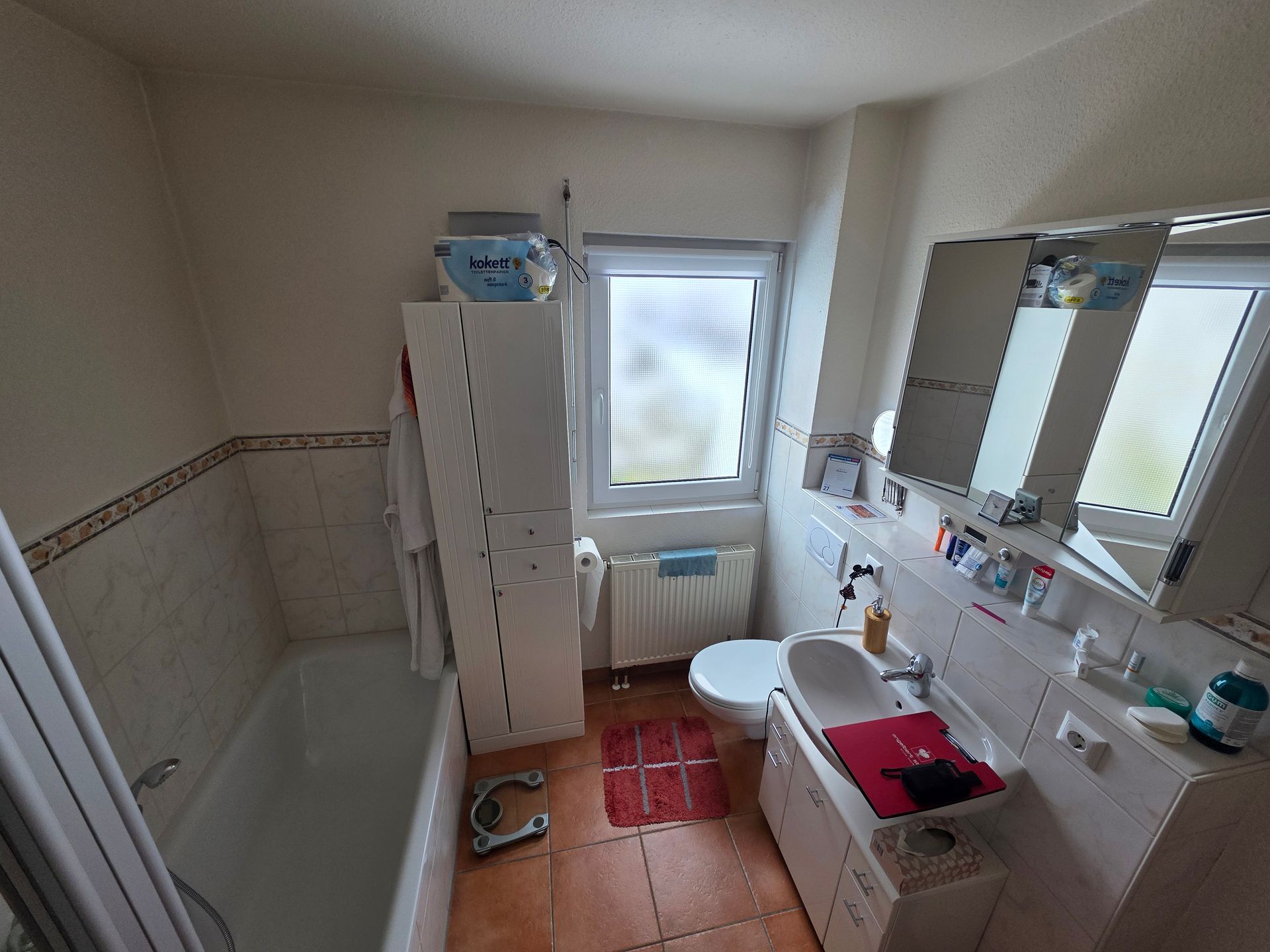 Kleines Badezimmer mit Badewanne, Waschbecken, Toilette, Fenster, rotem Teppich und Toilettenartikeln auf der Ablage.