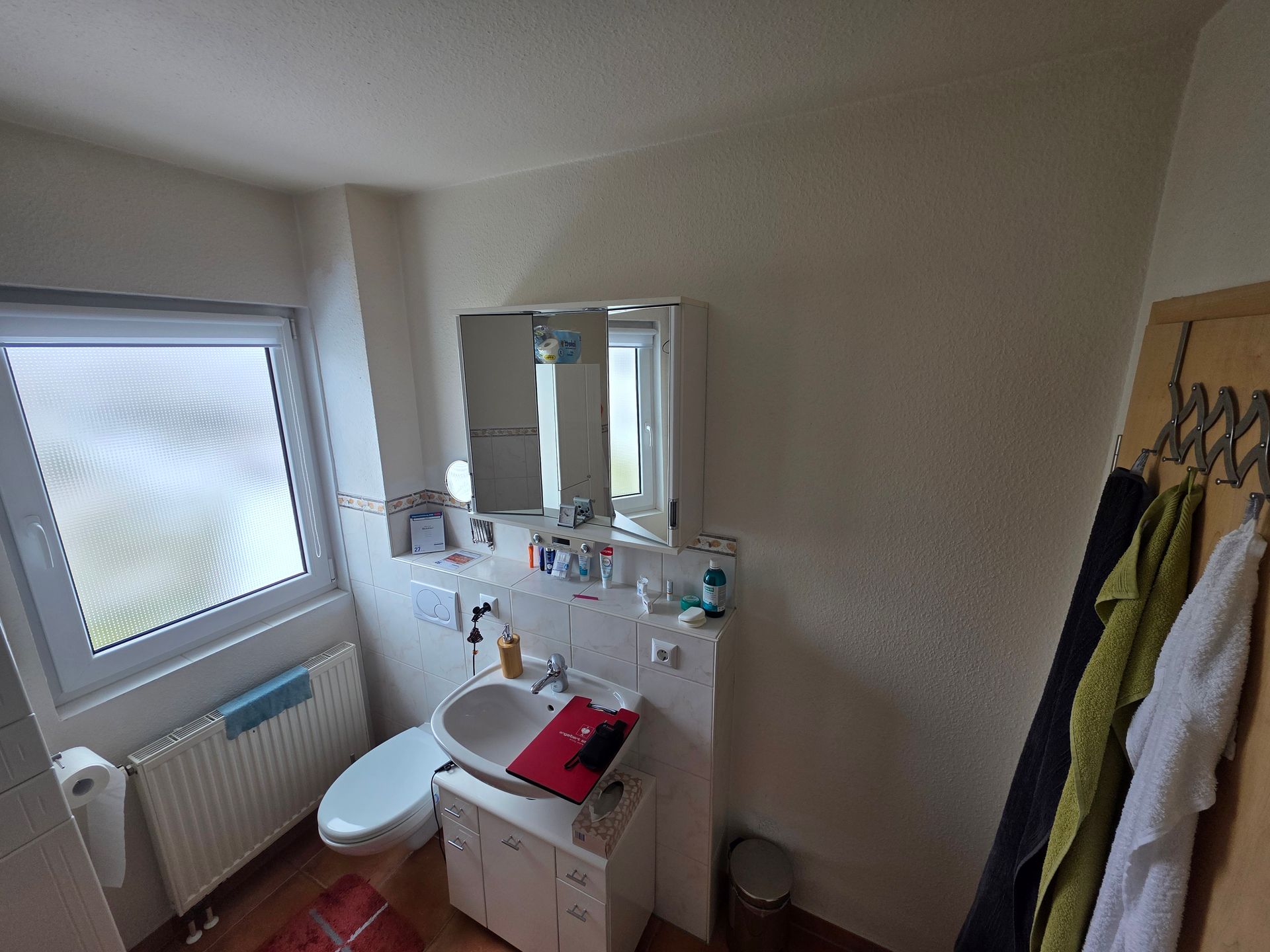 Kleines Badezimmer mit Waschbecken, Spiegel, Toilette, Milchglasfenster, Heizkörper und Bademantelhaken an der Wand.