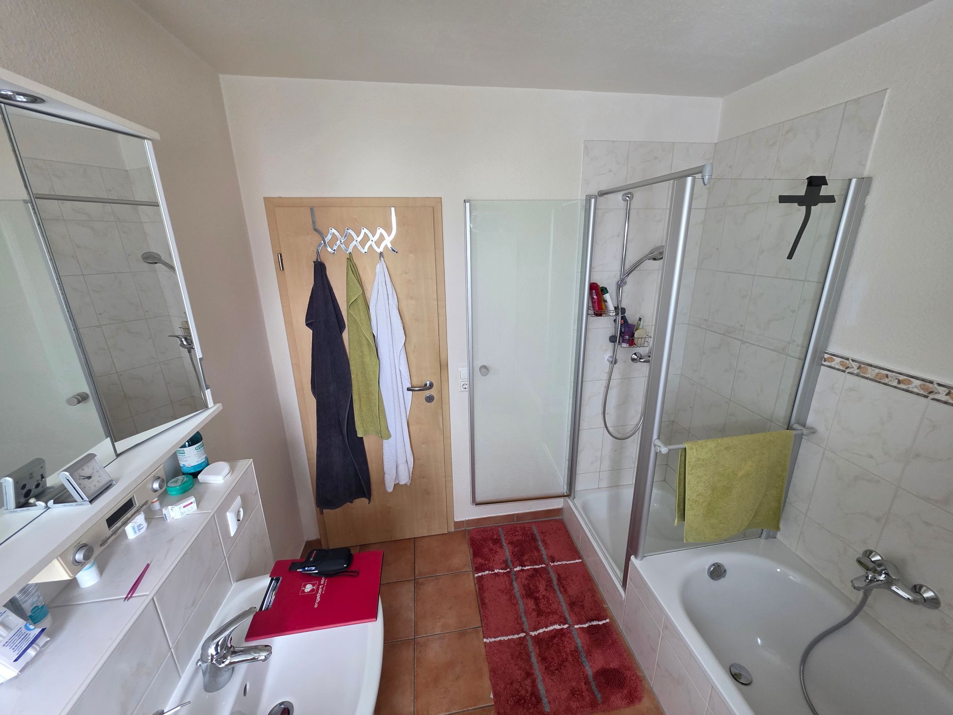 Badezimmer mit rotem Fliesenboden, weißer Badewanne mit Duschvorrichtung, Waschbecken, Spiegel und an der Wand befestigtem Kreuz.