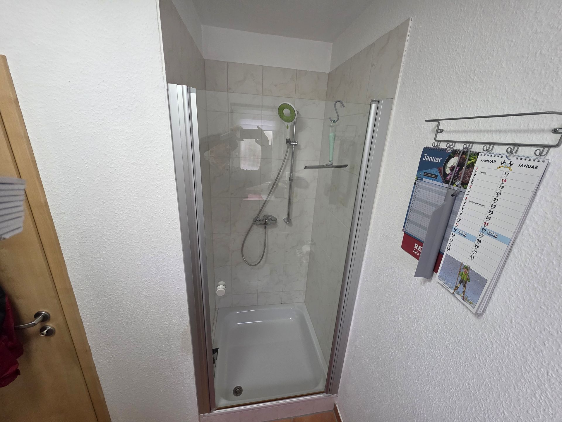 Kleine Duschkabine in einer schmalen Badezimmernische mit Handbrause und Wandkalender.