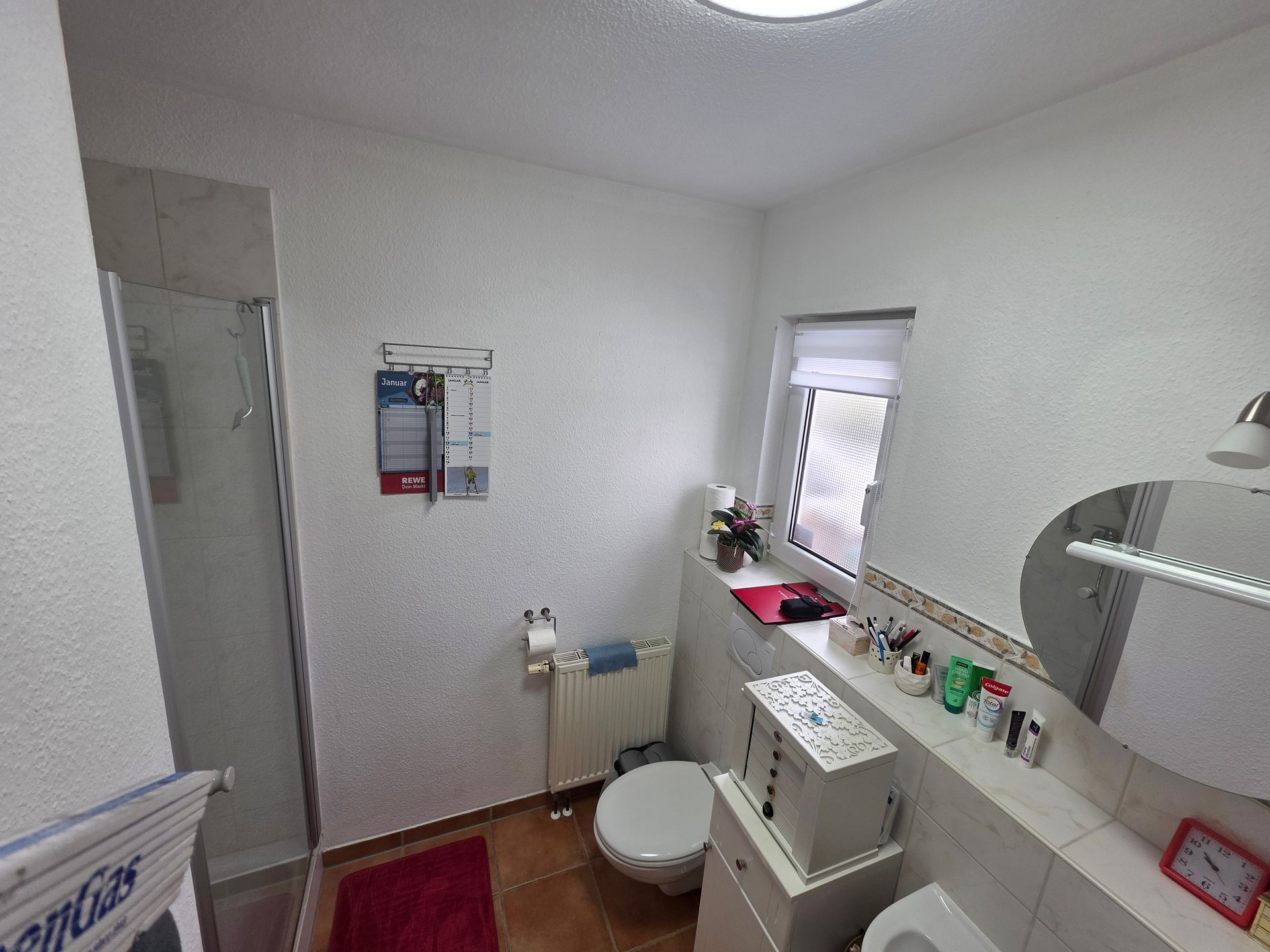 Kleines weißes Badezimmer mit Toilette, Waschbecken, Spiegel, Fenster und Reinigungsmitteln auf einem Regal.