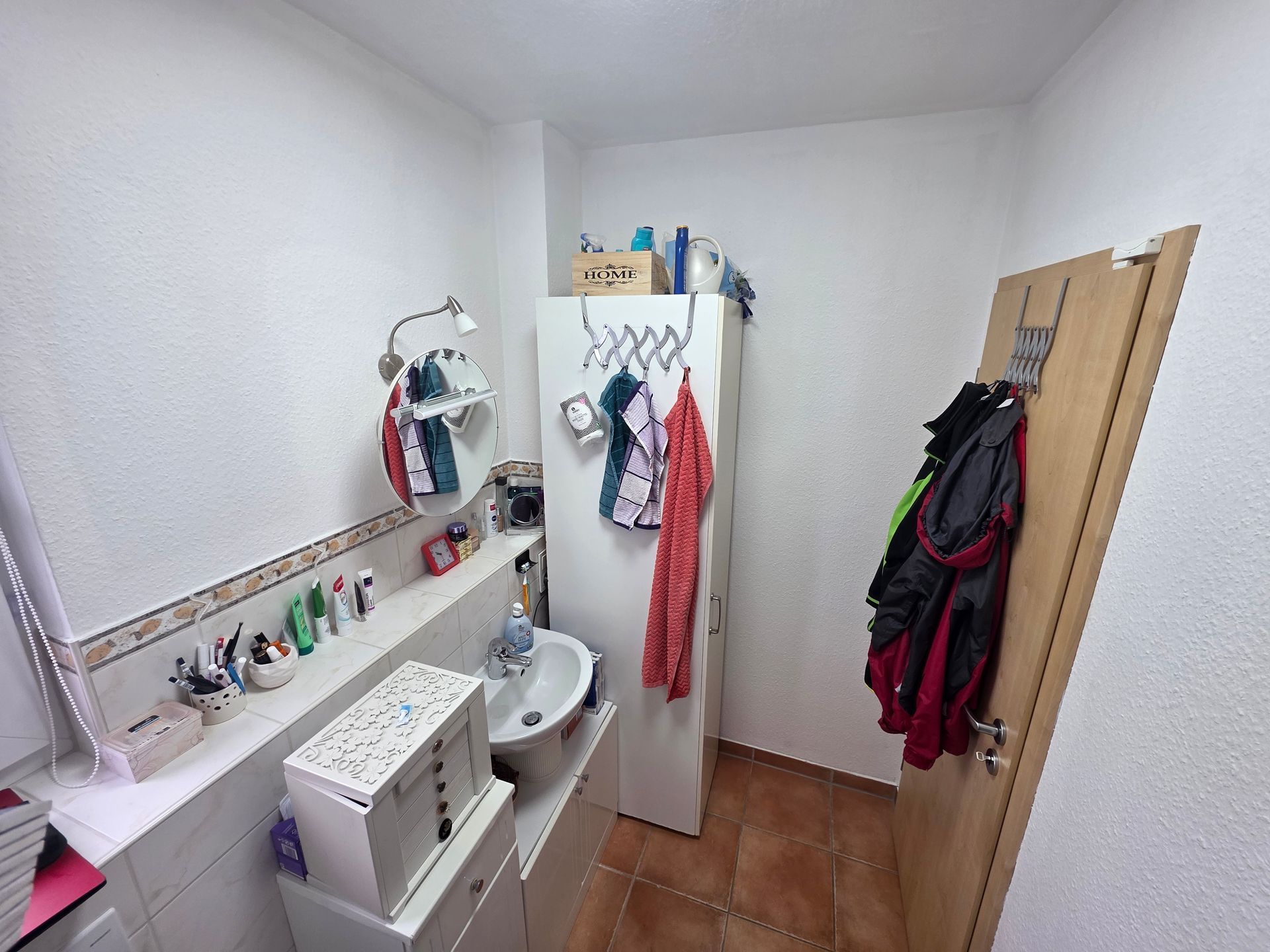 Kleines Badezimmer mit Waschbecken, Toilette, Wandhaken, Heizkörper und offener Tür, an deren Seite Mäntel hängen.