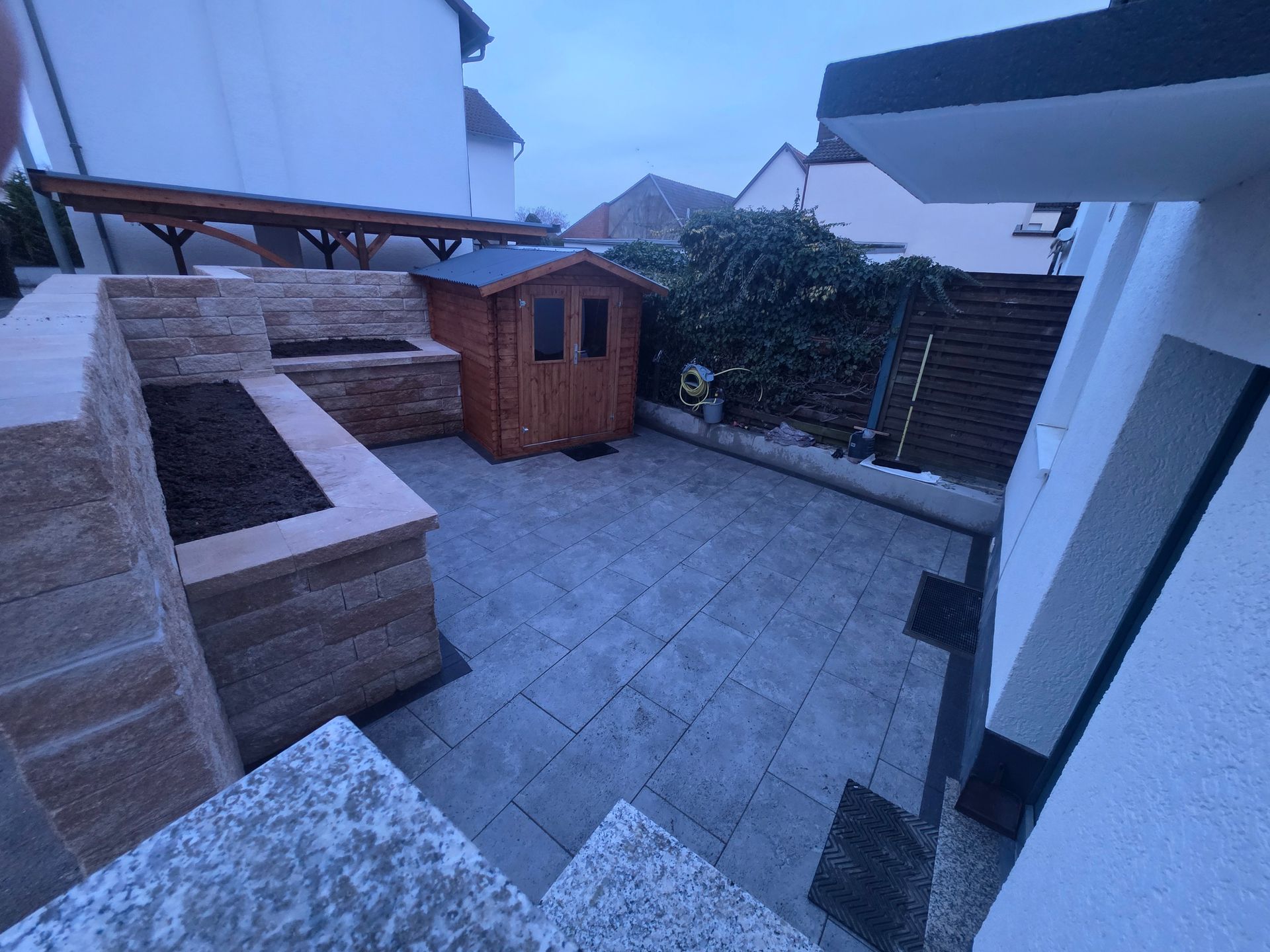 Steingepflasterte Terrasse im Hinterhof mit Holzschuppen, steinernen Pflanzbänken und Hecke entlang des Zauns in der Abenddämmerung