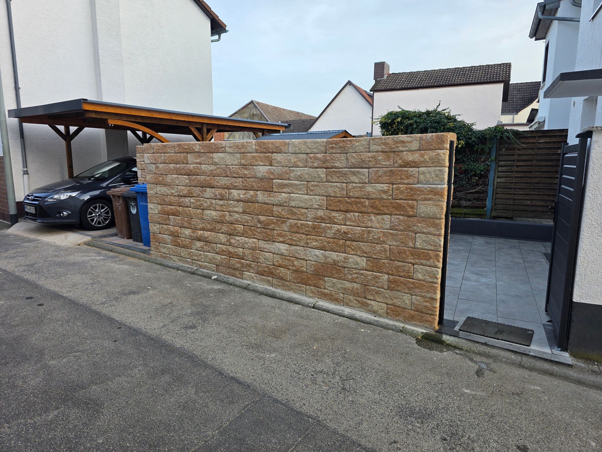 Steinmauer und ein Tor führen zu einer Einfahrt neben einer abfallenden Straße, unter einem Carport steht ein geparktes Auto.