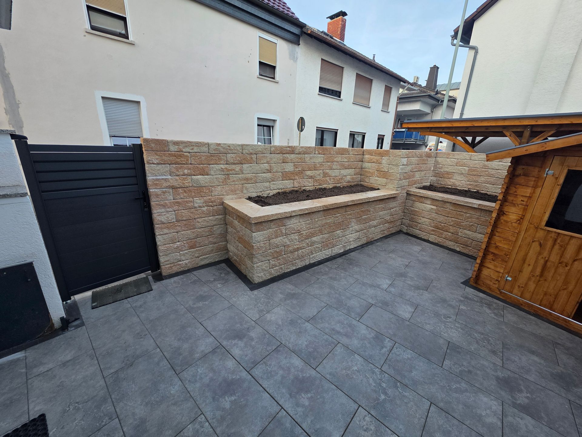 Terrasse mit grauen Pflastersteinen, steinernen Sitzmauern, schwarzem Tor und Holzschuppen neben Wohnhäusern