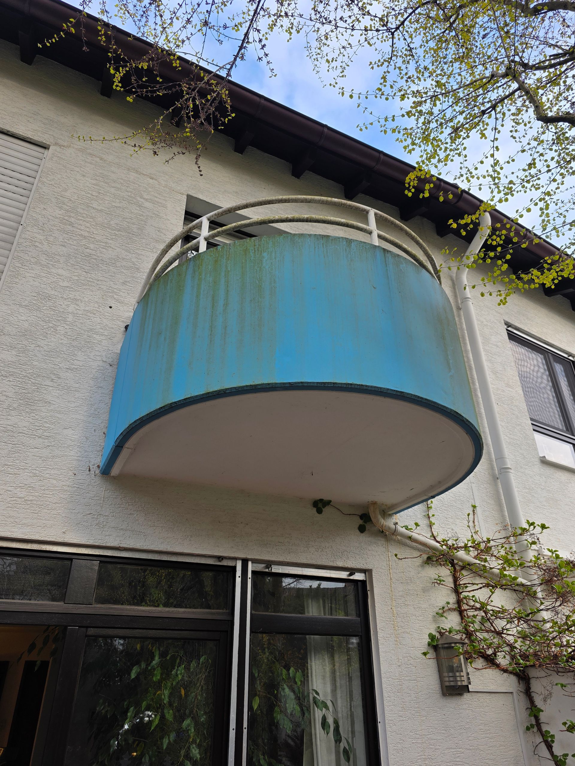 Blauer, geschwungener Balkon an der Gebäudeaußenseite, über einem Fenster und einer Tür, mit Grün entlang der Dachlinie
